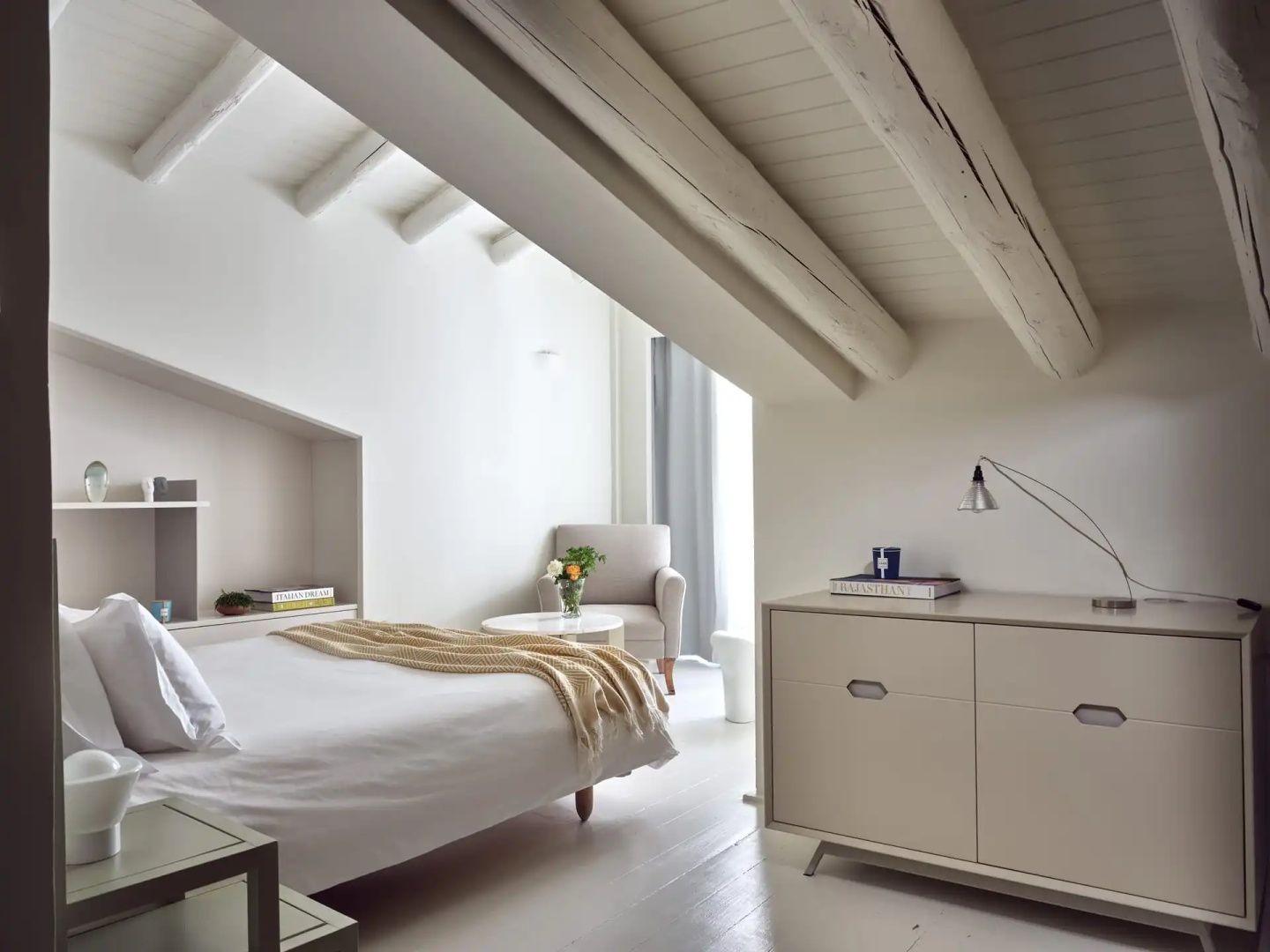 Helles Dachgeschosszimmer mit weißem Bett, Holzbalken an der Decke und einem Sessel.