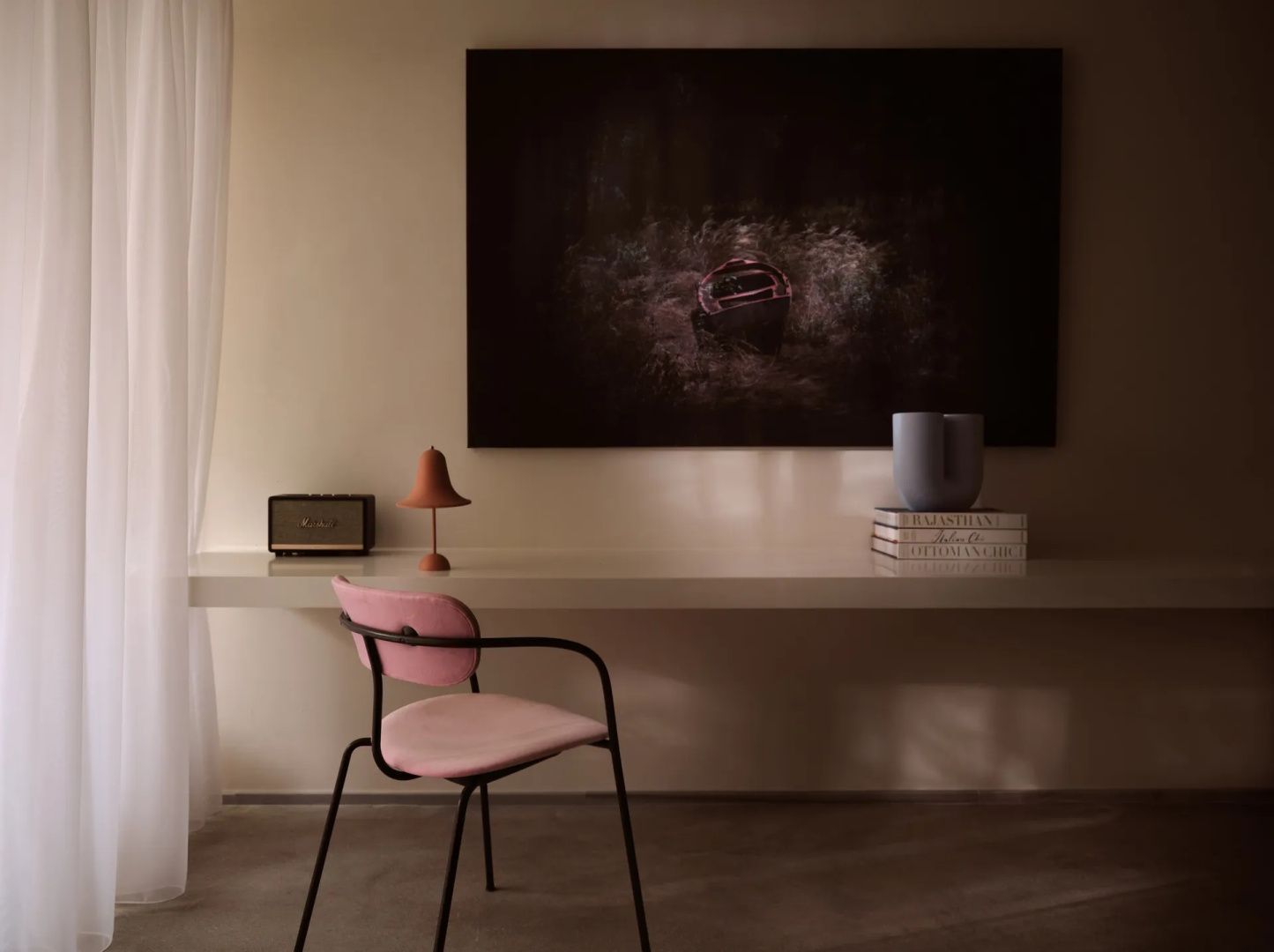 Schreibtisch mit einem rosa Stuhl, Tischleuchte und einem dunklen Wandbild.
