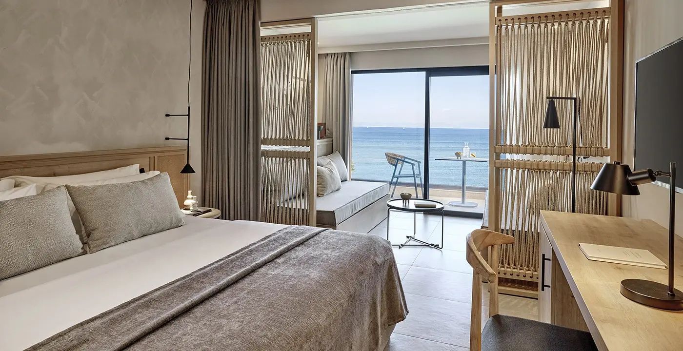Helles Hotelzimmer mit Doppelbett, Sitzbereich und Balkon mit Blick auf das Meer.