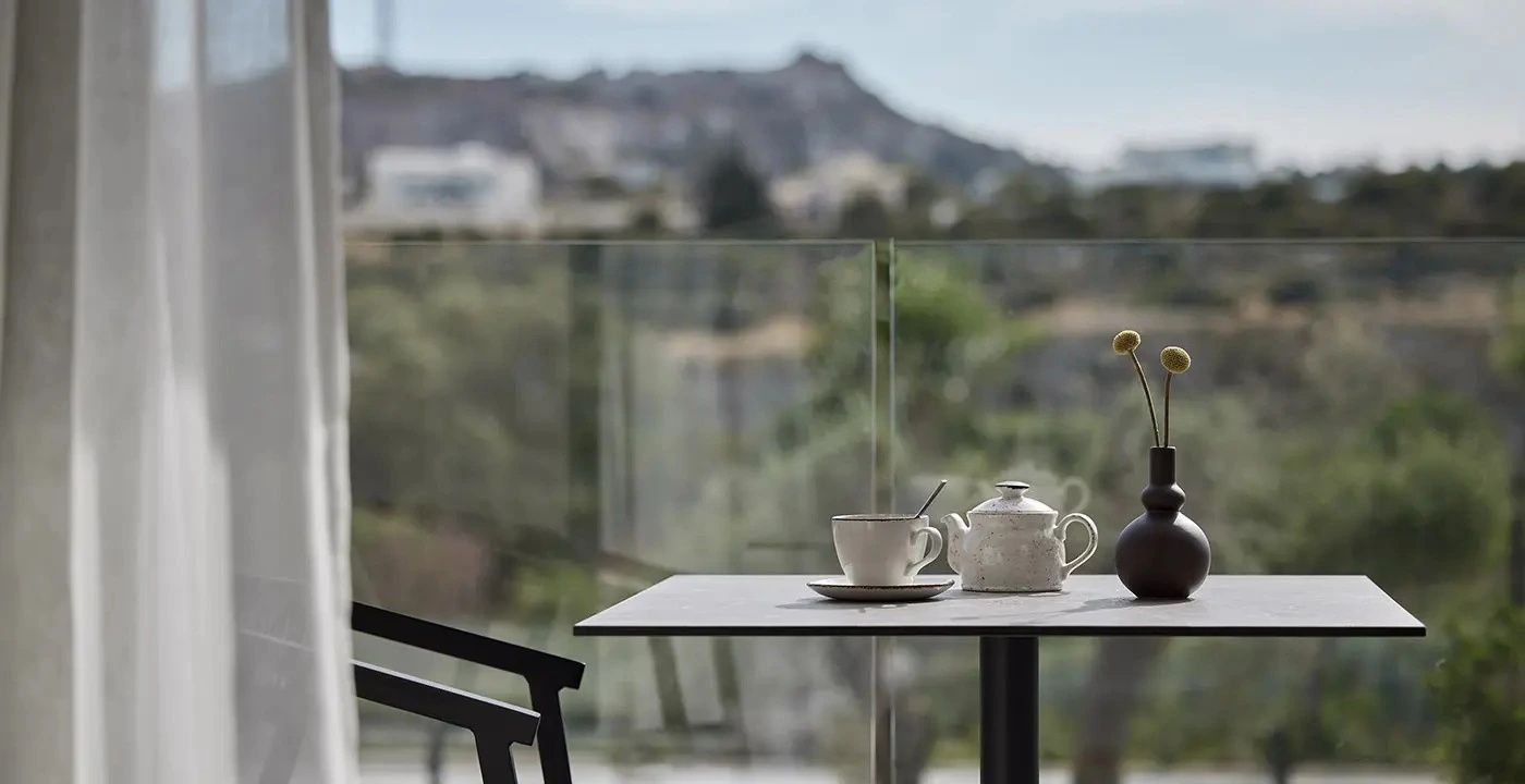 Gedeckter Tisch mit Teekanne auf einem Balkon mit Glasgeländer und Blick auf die Hügel.