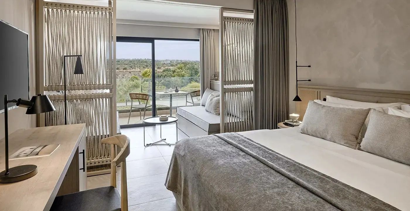 Modernes Hotelzimmer mit hellen Möbeln, Raumteiler und Balkon mit Blick ins Grüne.