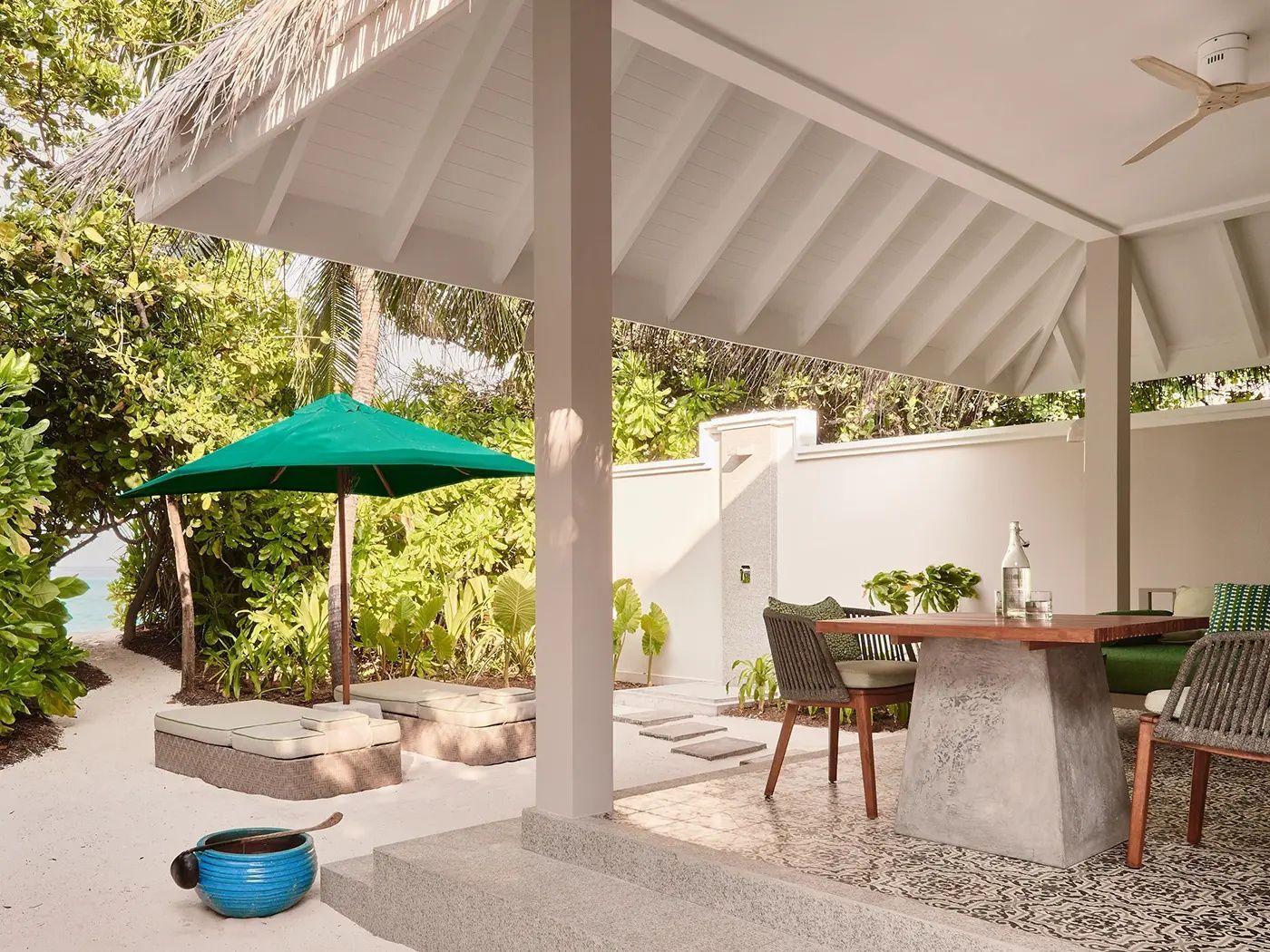 Überdachte Terrasse mit Esstisch, Sonnenliegen und grünem Sonnenschirm im Außenbereich.