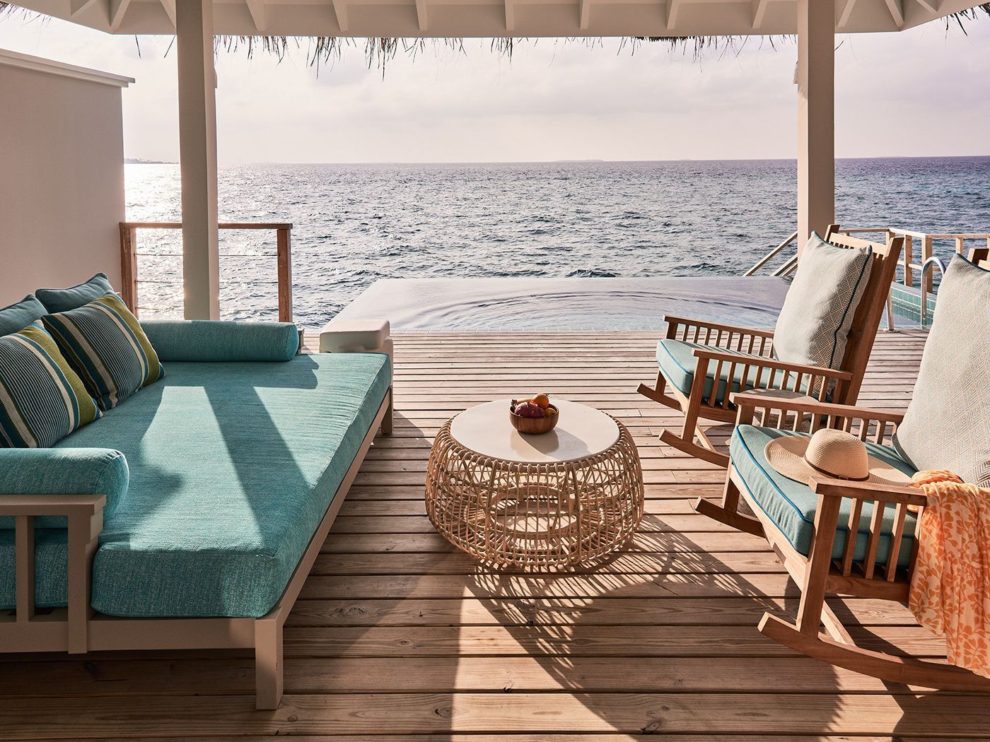 Holzterrasse einer Wasservilla mit türkisfarbenem Liegesofa, Schaukelstühlen, Pool und Blick auf den Ozean.