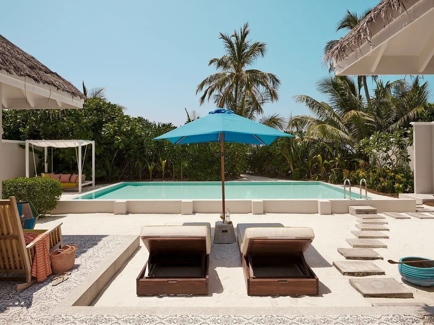 Zwei Holzsonnenliegen mit blauem Sonnenschirm an einem Pool vor Palmen.