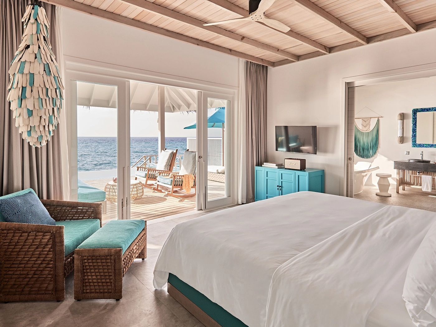 Schlafzimmer einer Wasservilla mit Doppelbett, blauen Möbeln und Blick auf die Terrasse und das Meer.