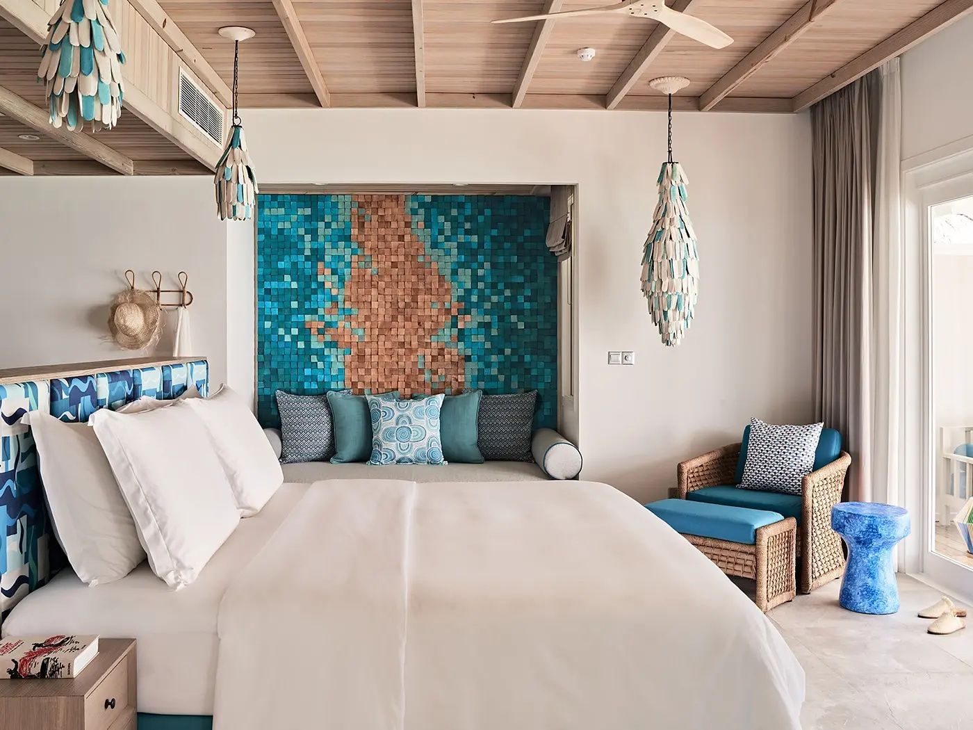 Schlafbereich mit weißem Doppelbett, blauer Mosaikwand und Sessel aus Rattan.