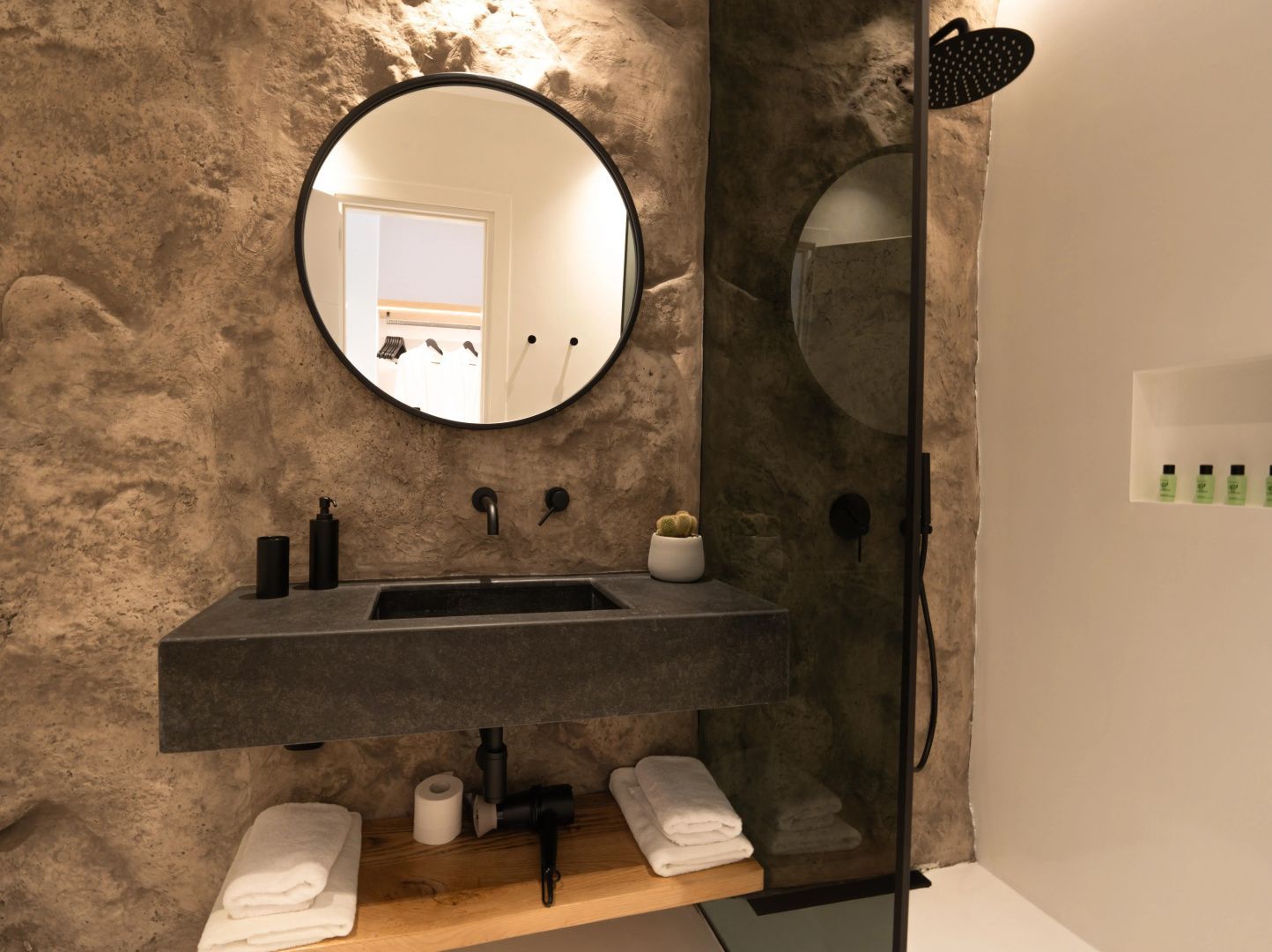 Badezimmer mit dunklem Waschtisch, rundem Spiegel und Steinwand sowie Dusche.