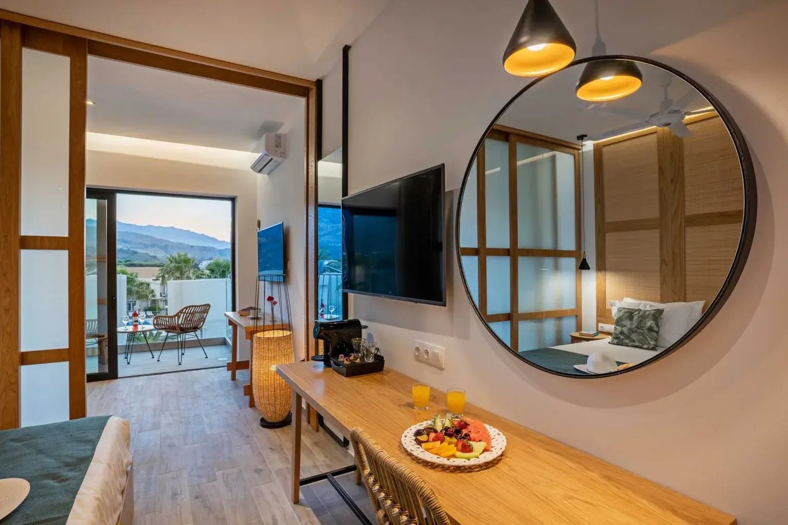 Schreibtisch mit Fernseher und rundem Spiegel in einem hellen Hotelzimmer mit Balkon.