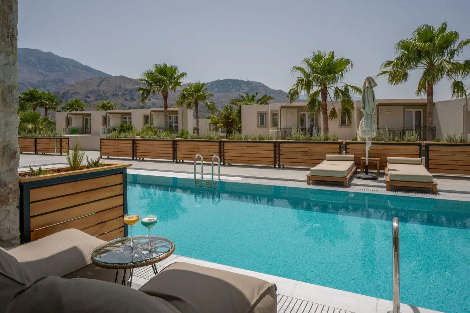 Poolbereich mit Sonnenschirm, Liegen und Palmen vor flachen Hotelgebäuden und Bergen.