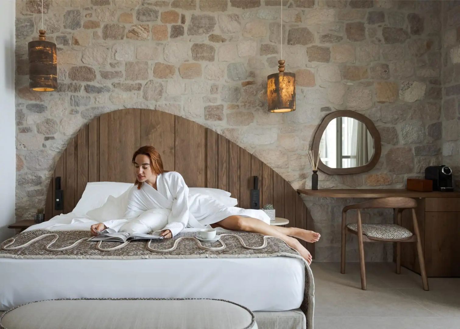 Frau im Bademantel liest ein Magazin auf dem Bett in einem Zimmer mit Steinwand.