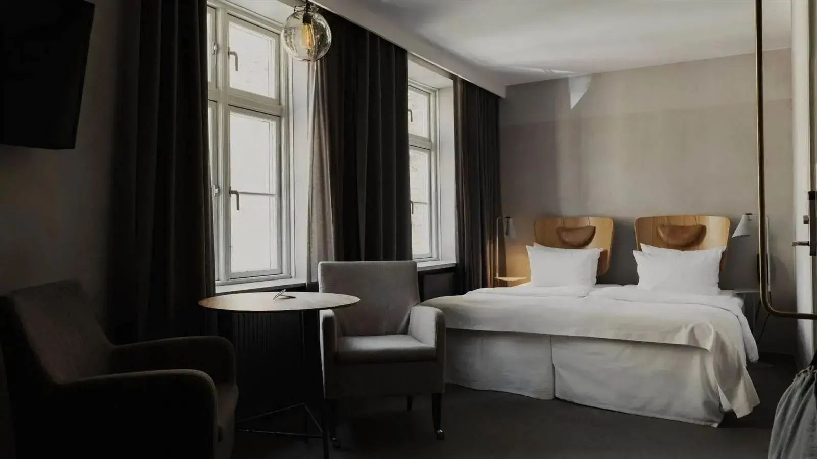 Helles Hotelzimmer mit Doppelbett, Sessel, Tisch und hohen Fenstern.