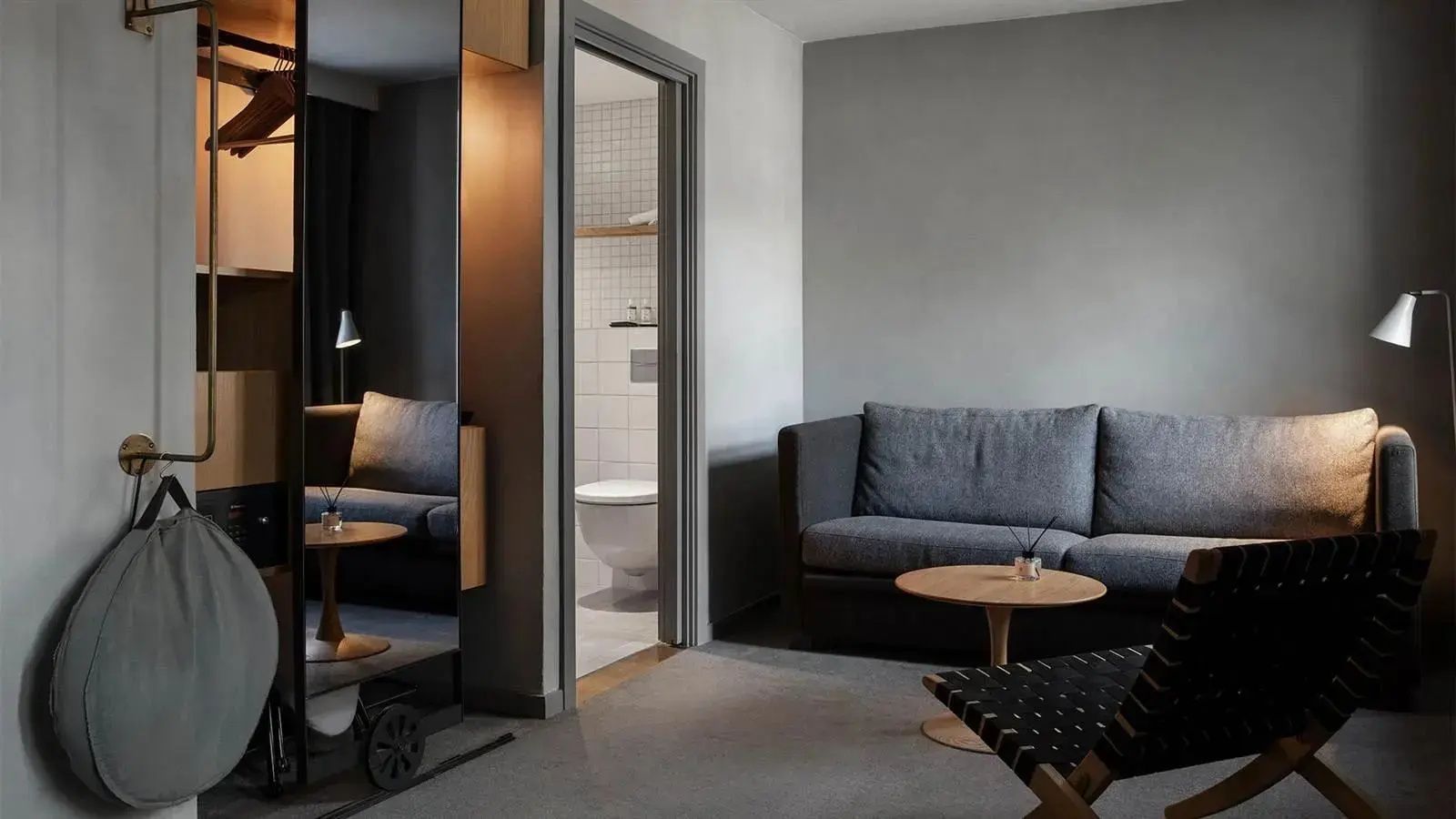 Wohnbereich eines Hotelzimmers mit grauem Sofa, Tisch und Blick ins Badezimmer.
