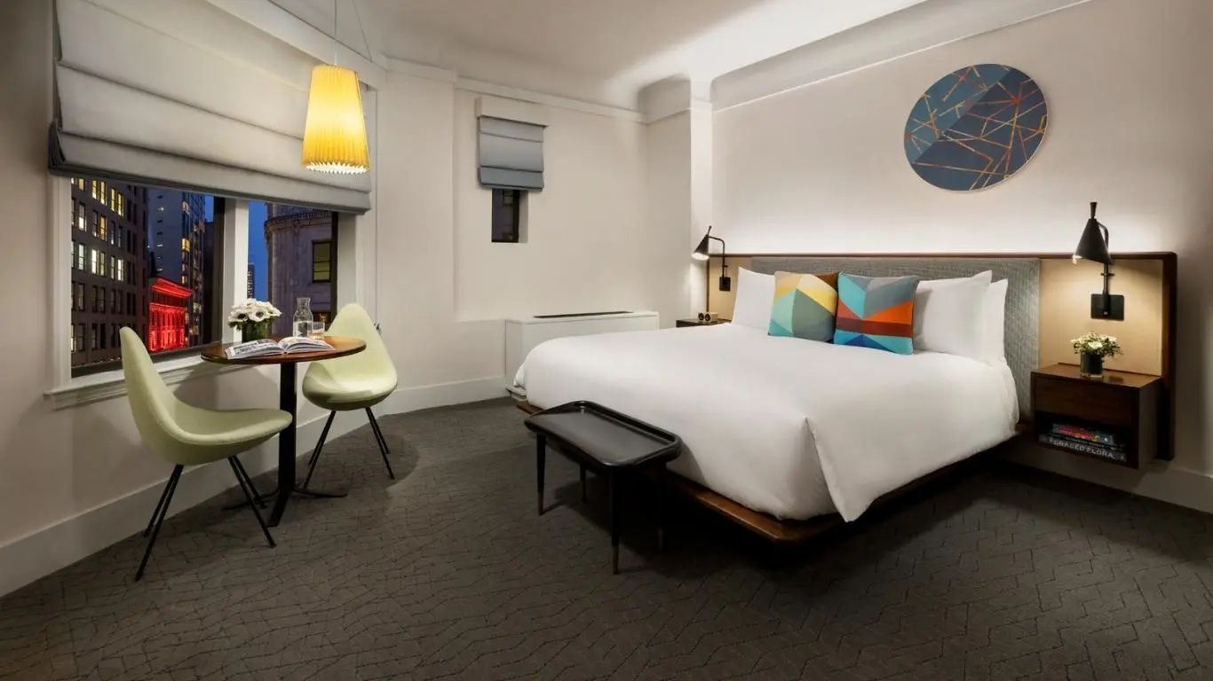 Hotelzimmer mit großem Bett, bunten Kissen, kleiner Sitzbank und Tisch mit zwei Stühlen am Fenster.