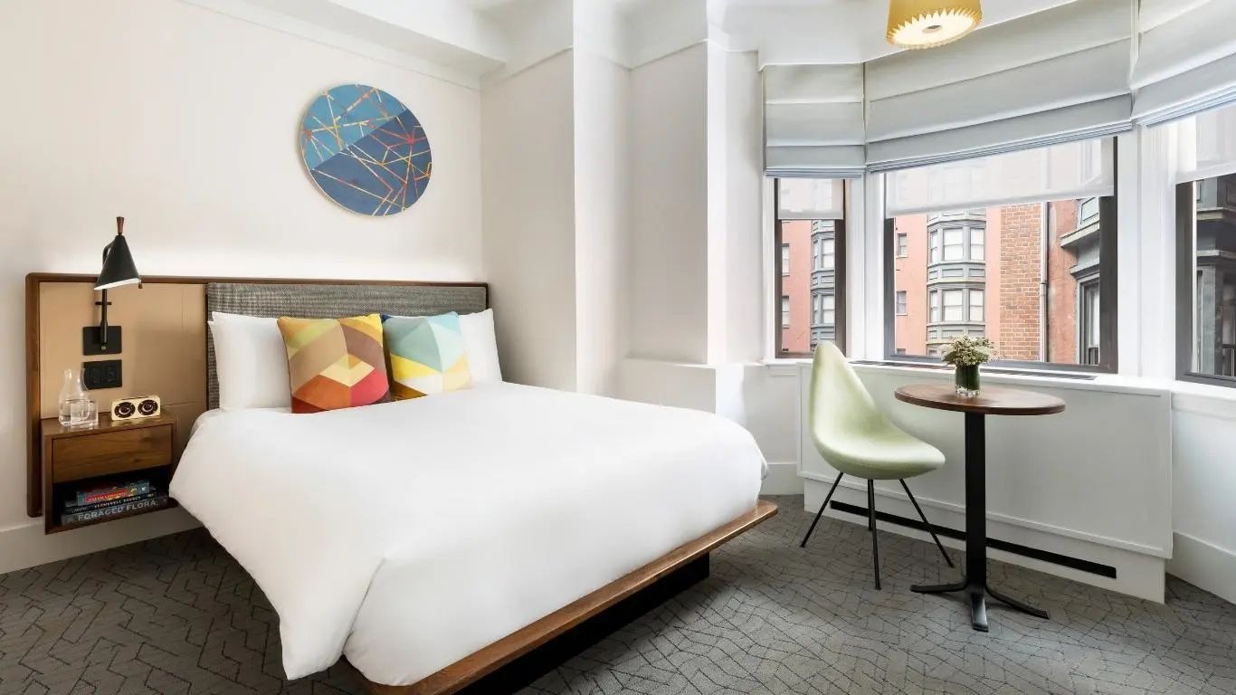Helles Zimmer mit Doppelbett, rundem Wandbild und Sitzplatz am Fenster.