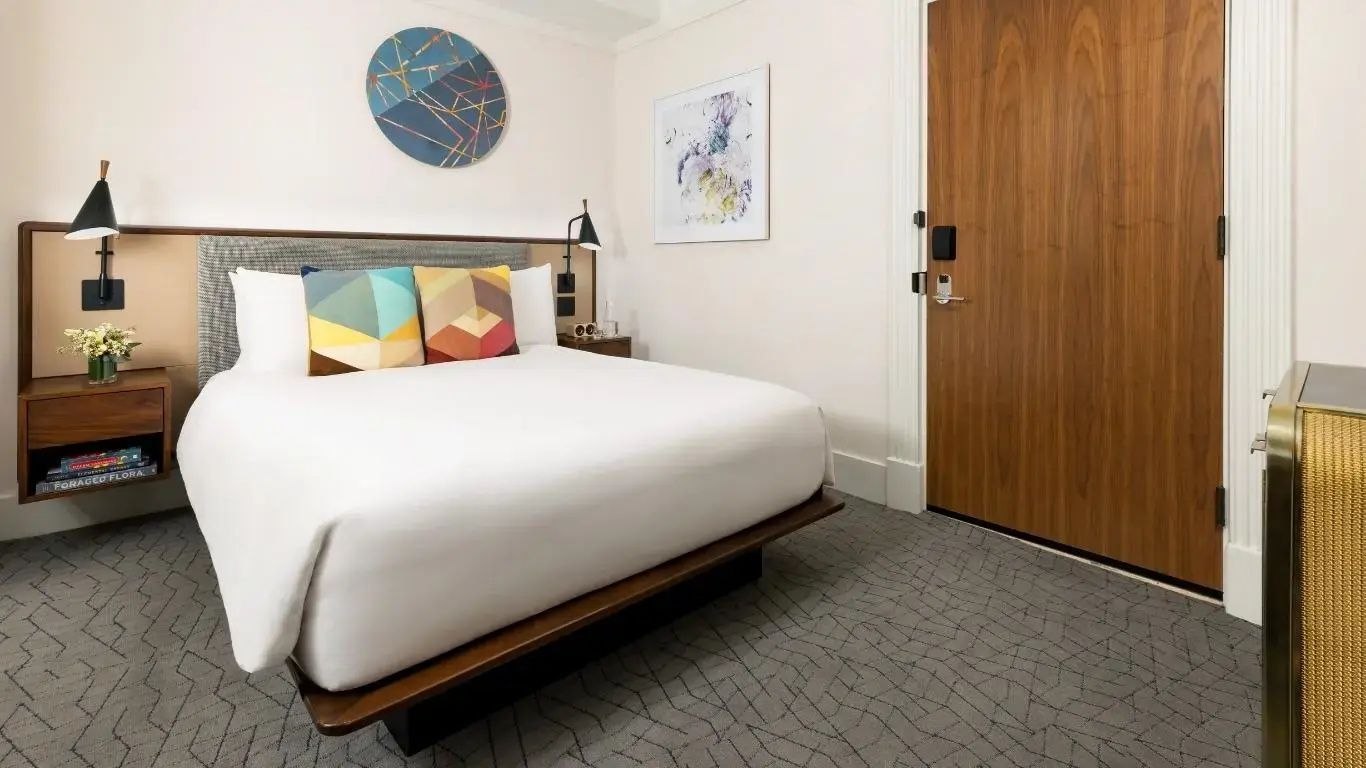 Hotelzimmer mit Doppelbett, bunten Dekokissen, Kunstwerk und Teppichboden.