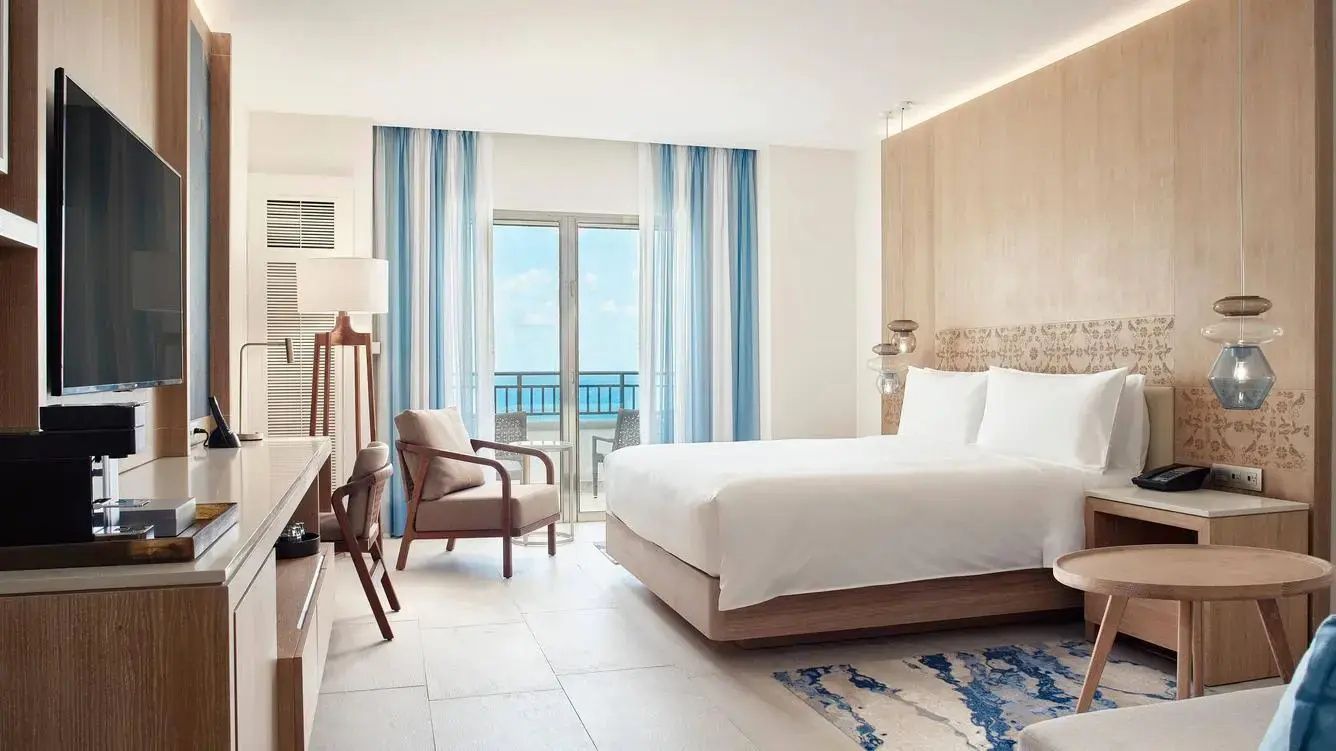 Helles Hotelzimmer mit Doppelbett, Sessel und Balkontür mit Blick auf das blaue Meer.