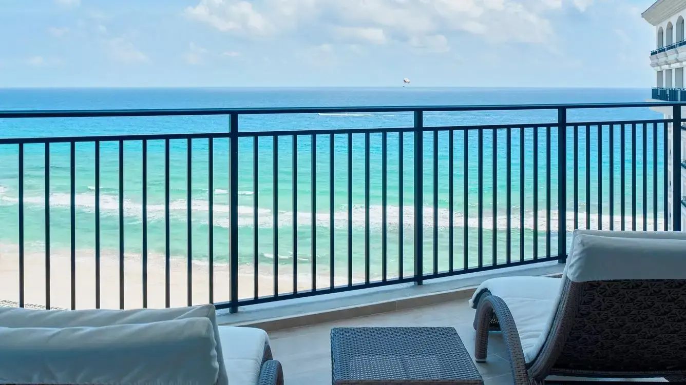 Blick vom Balkon über ein dunkles Geländer auf das türkisfarbene Meer und den Sandstrand.