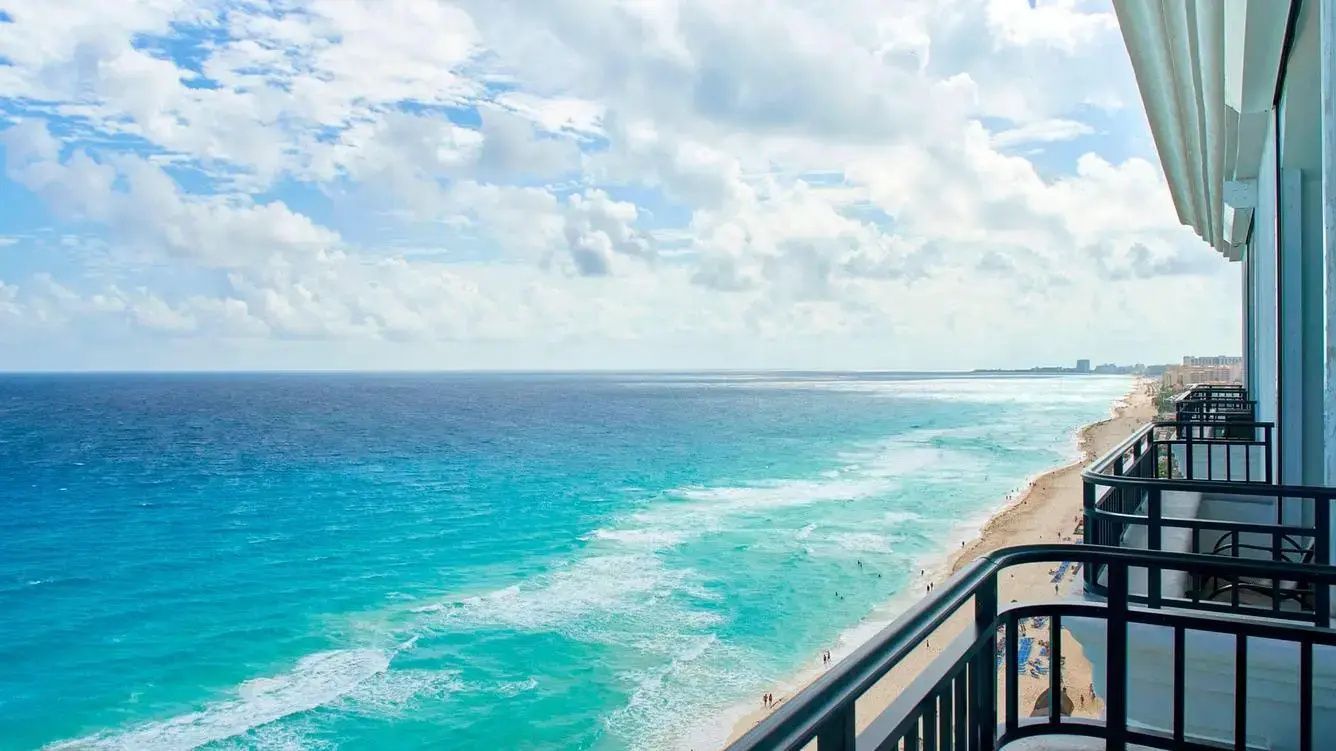 Blick von einem Balkon auf das türkisfarbene Meer und den feinen Sandstrand unter leicht bewölktem Himmel.