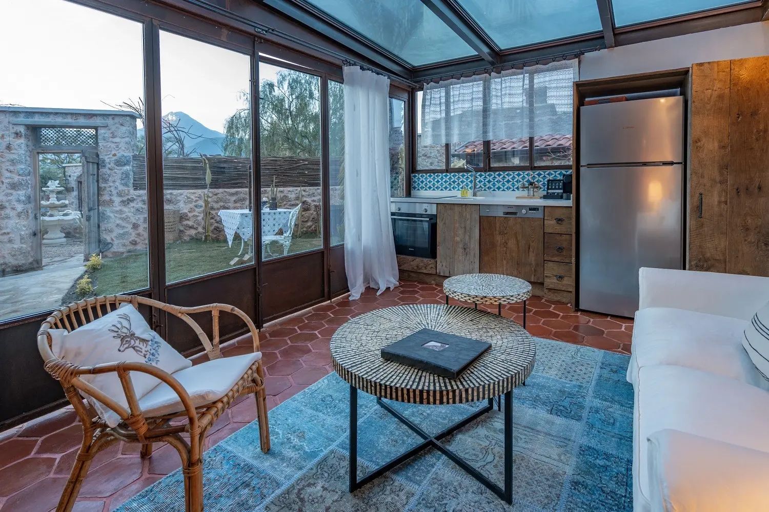 Verglaster Wohnbereich mit Küchenzeile, zwei Couchtischen, blauem Teppich und Blick in den Garten.