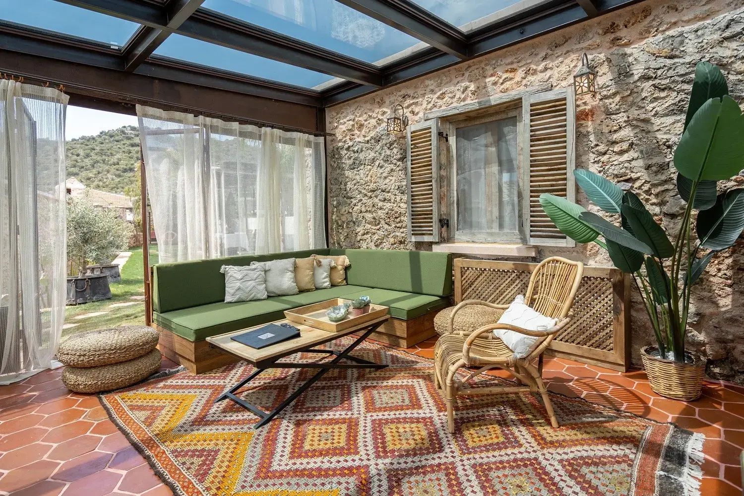 Überdachter Loungebereich mit grünem Ecksofa, Rattansessel und bunt gemustertem Teppich.