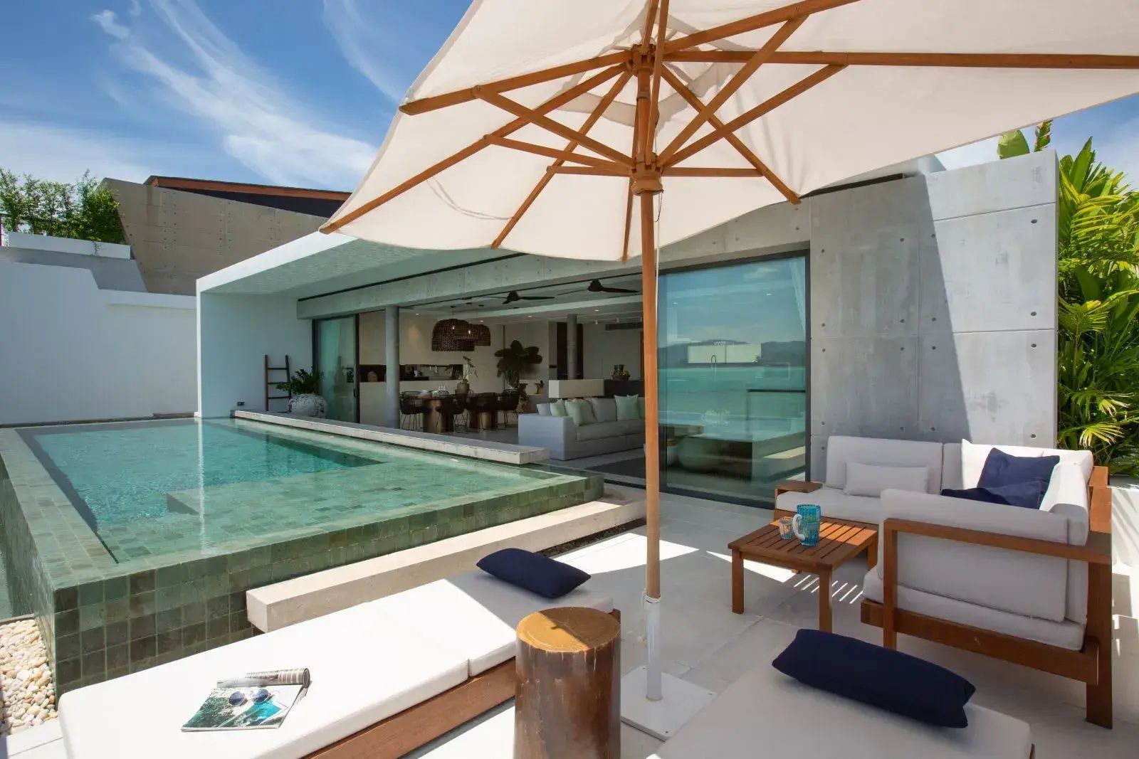 Sonnendeck mit Pool, Sonnenschirm und Sitzgelegenheiten vor dem geöffneten Wohnbereich.