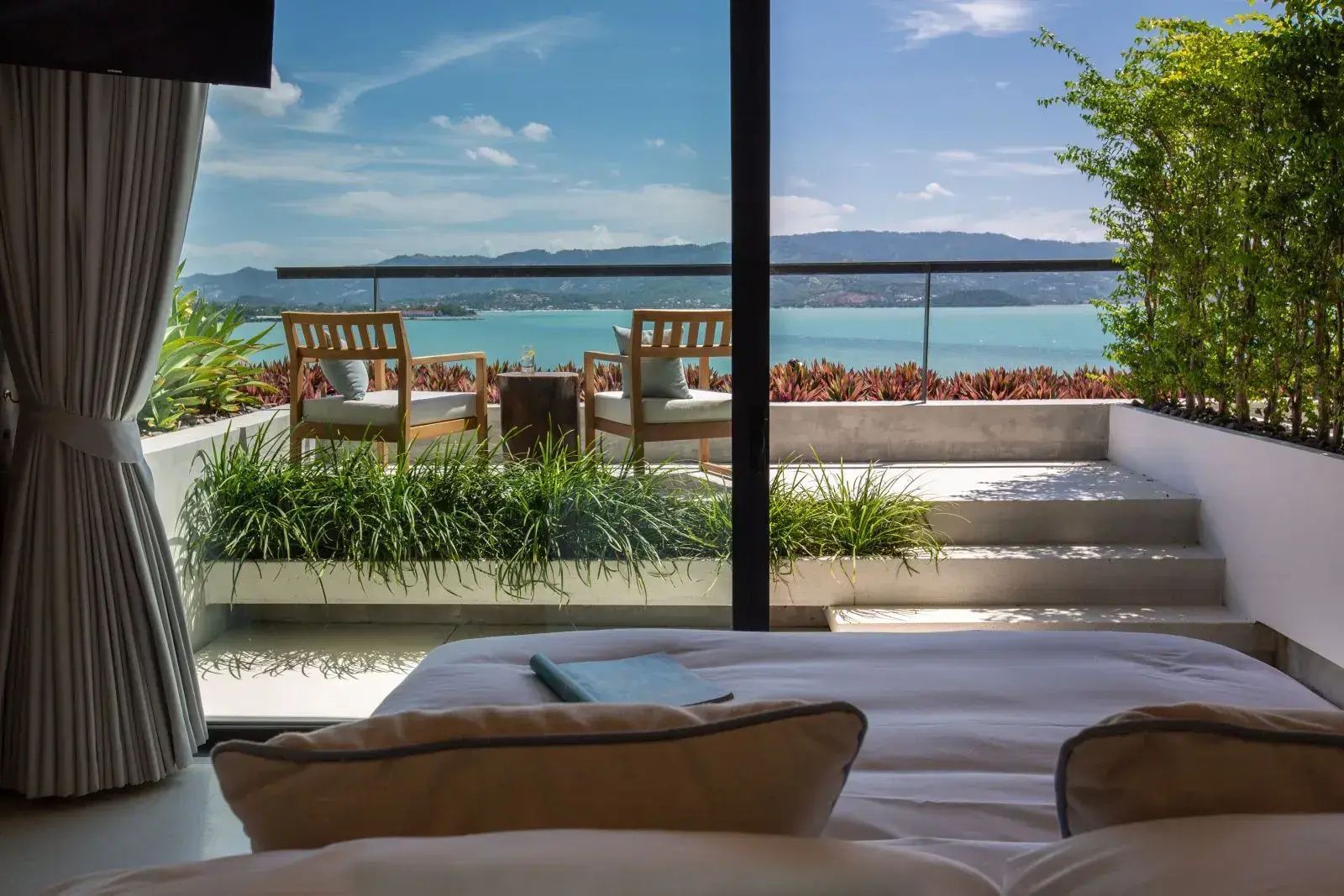Blick vom Bett durch eine Glasfront auf die Terrasse mit Stühlen und Meerpanorama