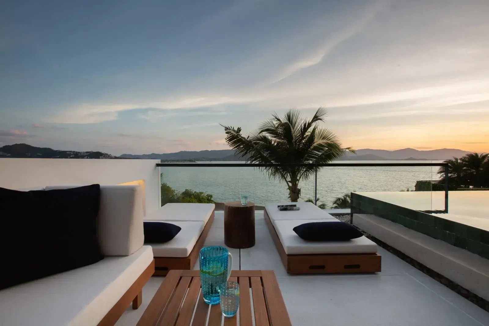 Terrasse mit braunen Sonnenliegen und Holztisch mit Blick auf das Meer im Abendlicht.