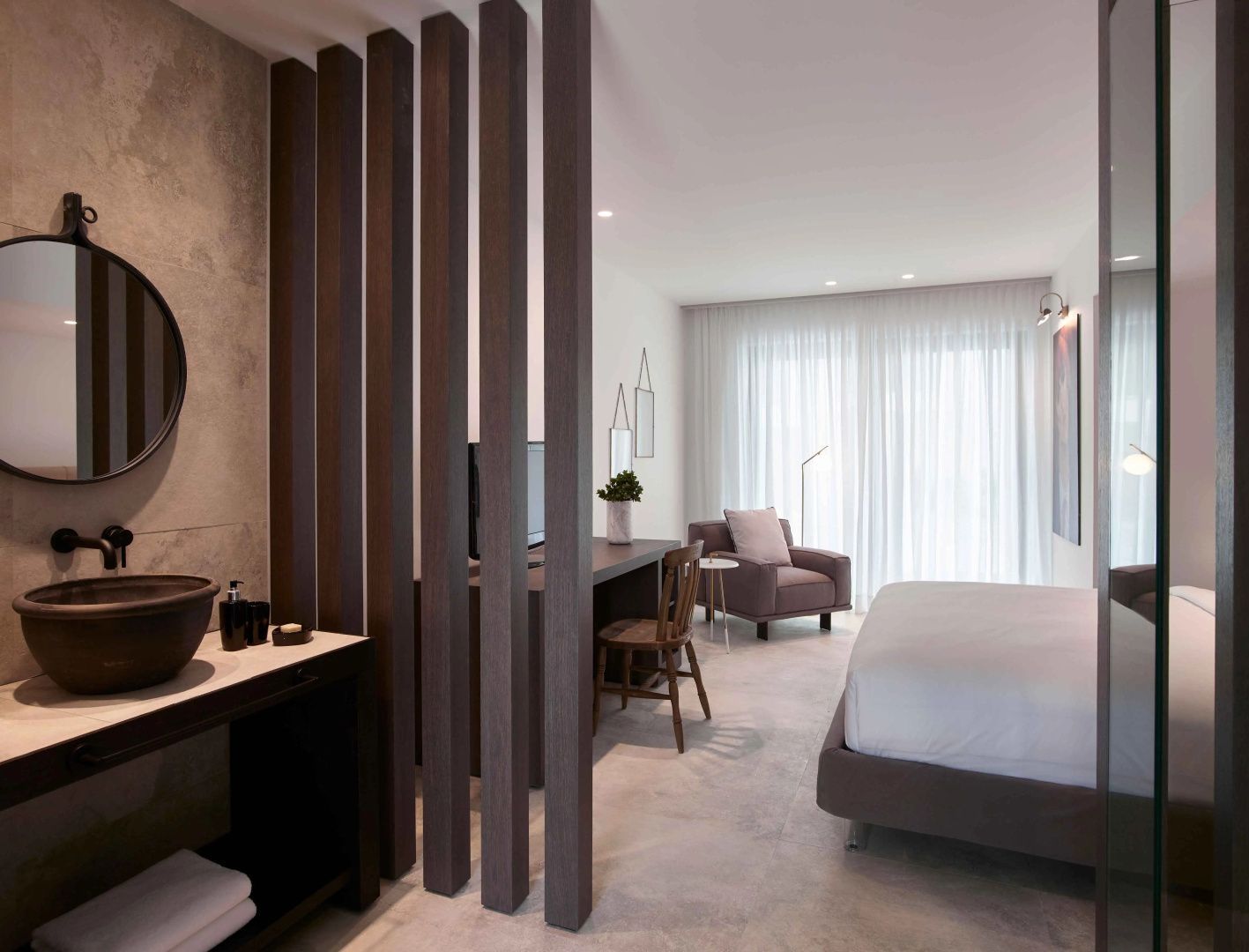 Offenes Hotelzimmer mit Waschtisch hinter Lamellenwand, Schreibtisch und Doppelbett.
