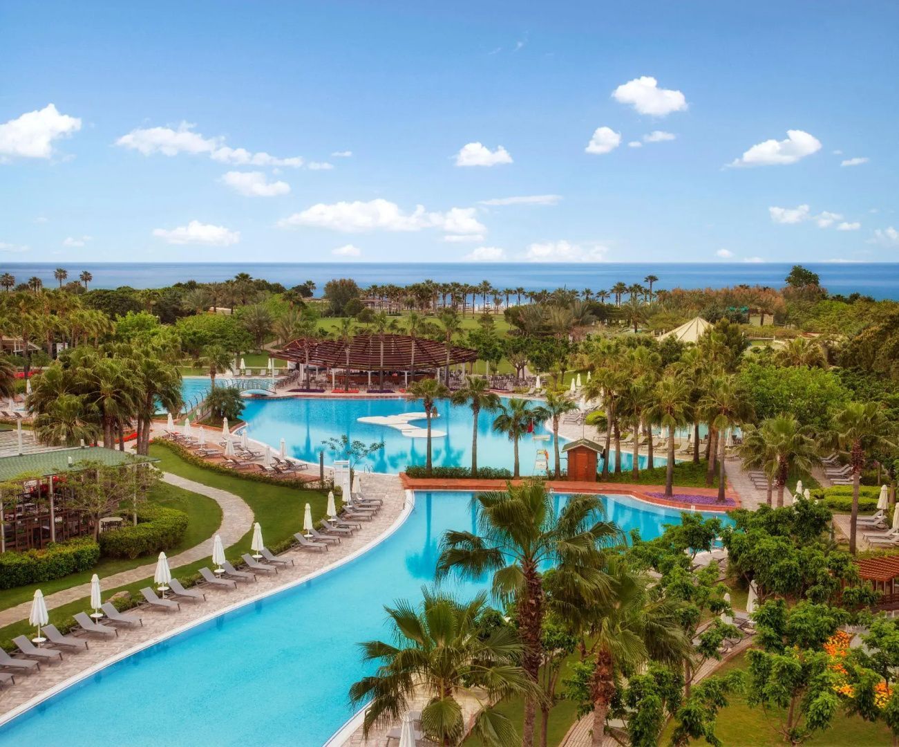 Große Poollandschaft im Hotelgarten mit Palmen und Blick auf das nahe Meer.