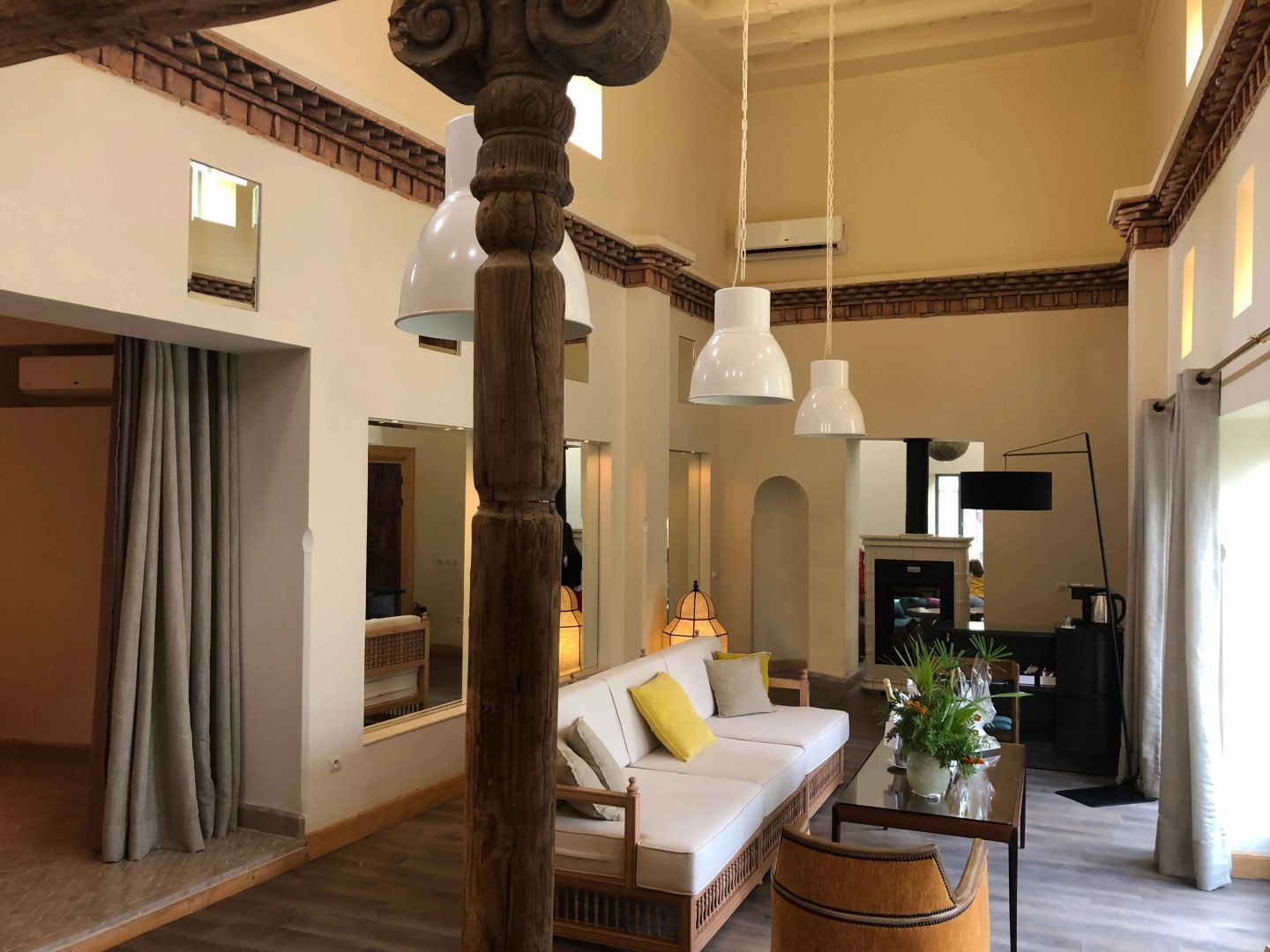 Sitzbereich mit weißer Couch, gelbem Kissen, Holzsäule und drei hängenden Lampen in einem hohen Raum.