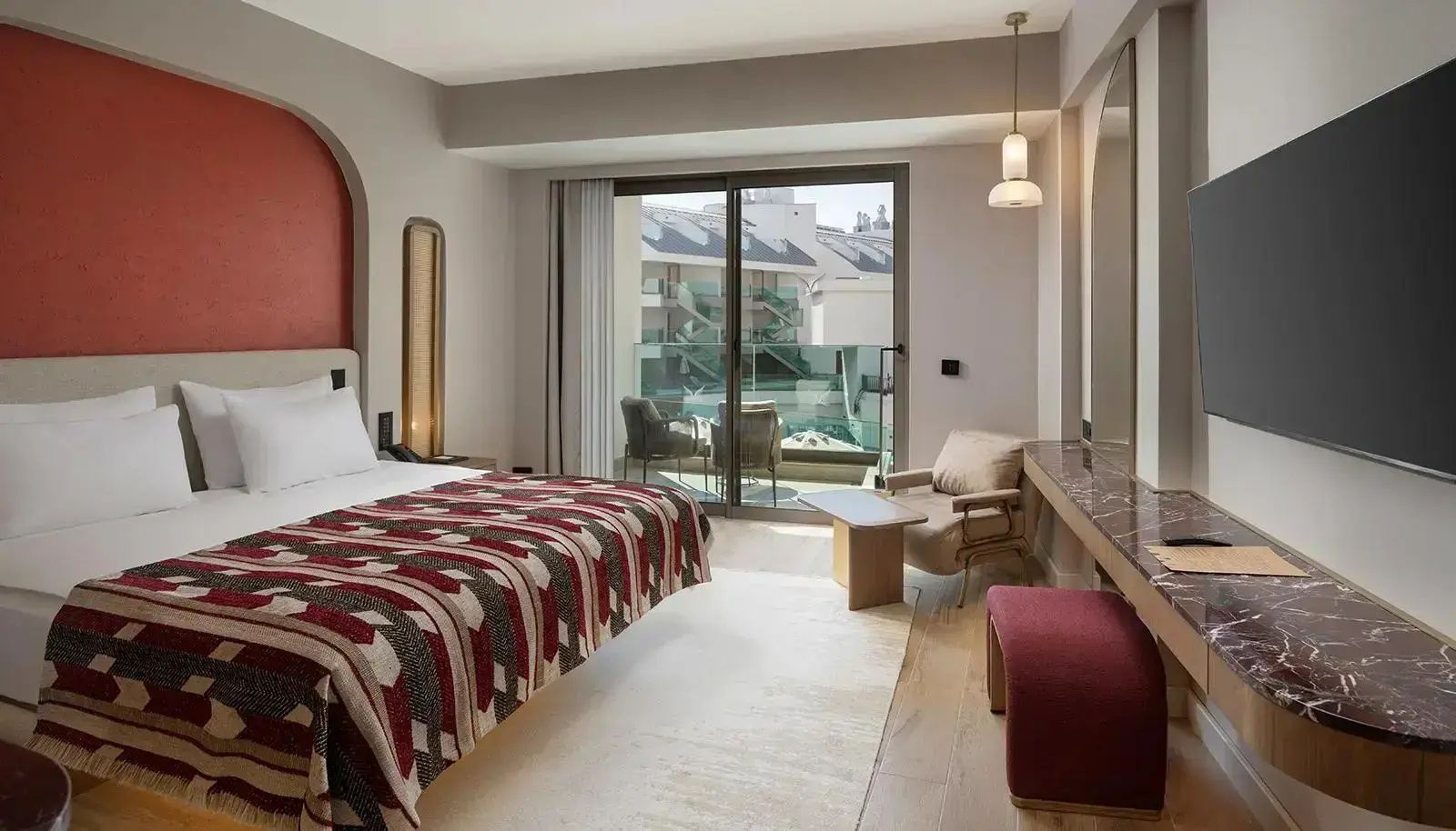 Helles Hotelzimmer mit rot gemusterter Tagesdecke, Sessel und Balkonzugang.