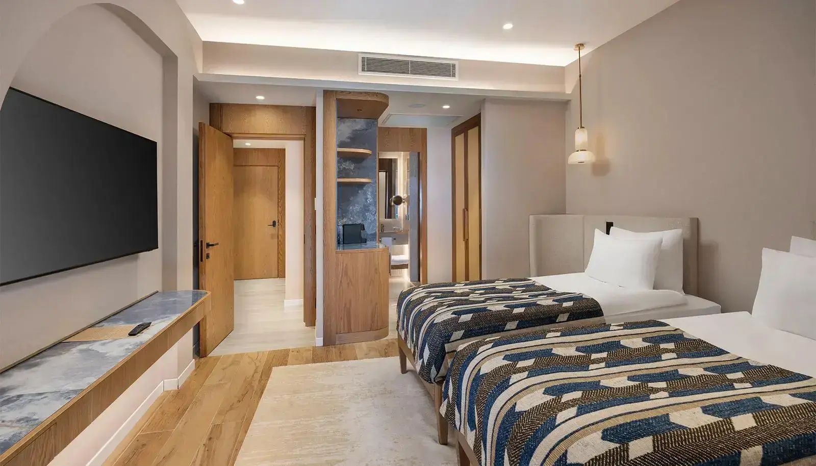 Hotelzimmer mit zwei Einzelbetten mit Musterdecken, Holzregal und Blick in den Flur.