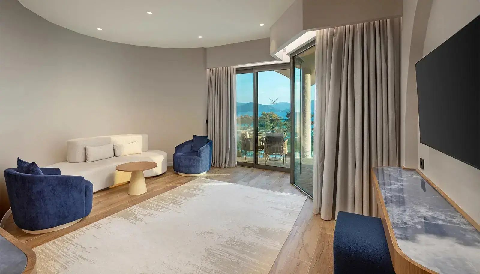 Wohnbereich des Zimmers mit weißem Sofa, blauem Sessel, Teppich und Panoramafenster.