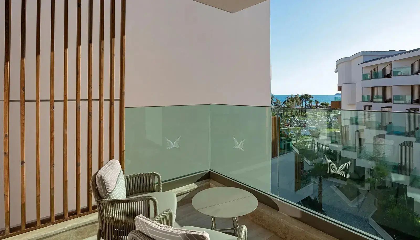 Balkon mit Sitzmöbeln, Glasbrüstung und Ausblick auf Palmen sowie das Meer.
