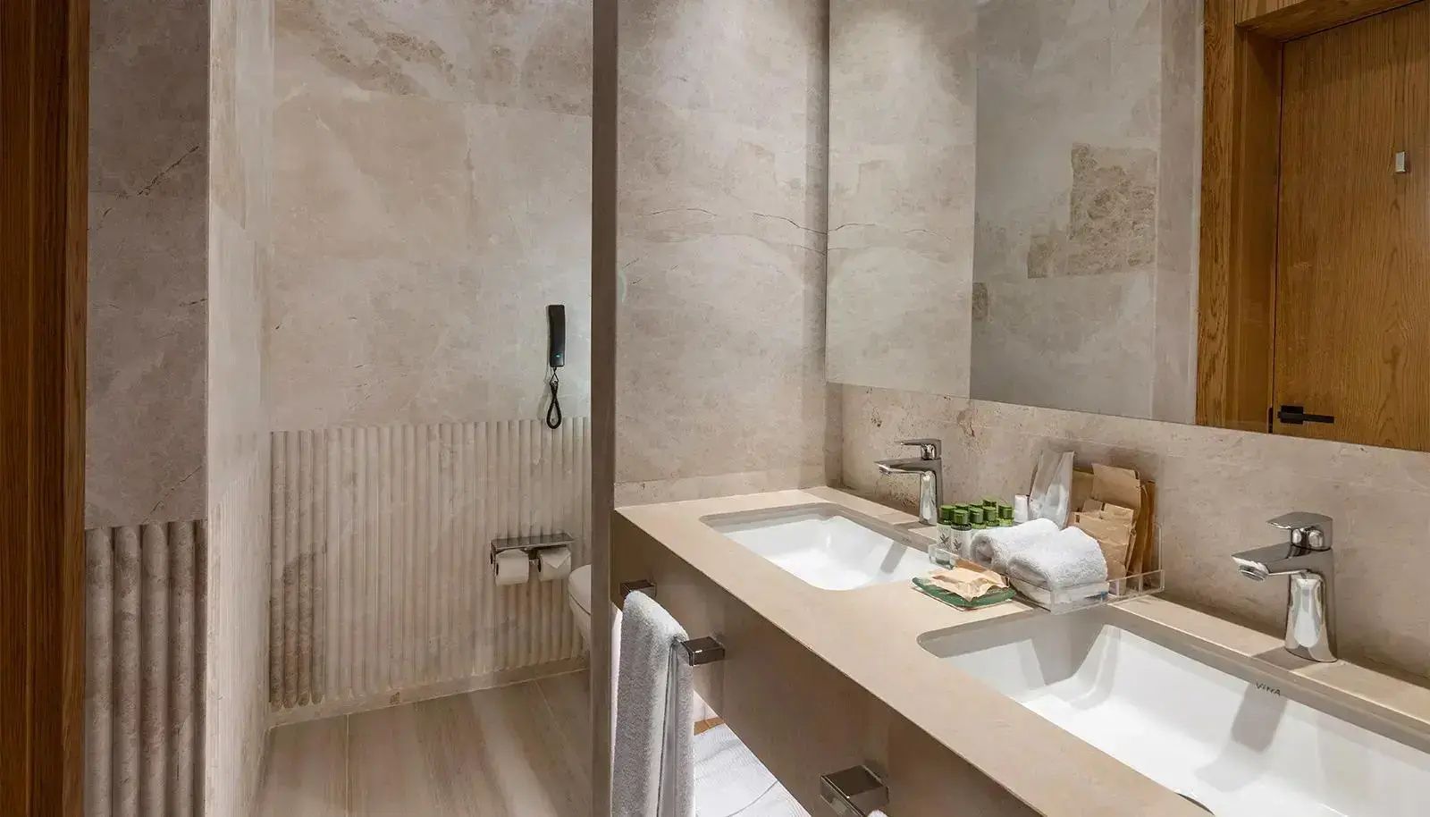 Badezimmer mit beige gefliestem Waschtisch, Spiegel, Handtüchern und Toilettenbereich.
