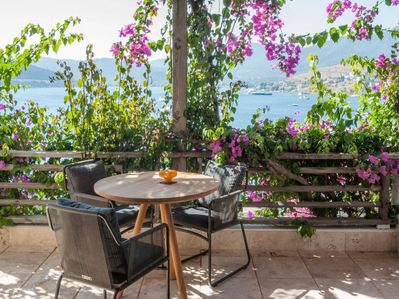 Überdachte Terrasse mit Holztisch, Stühlen, rosa Bougainvillea und Blick auf das blaue Meer.
