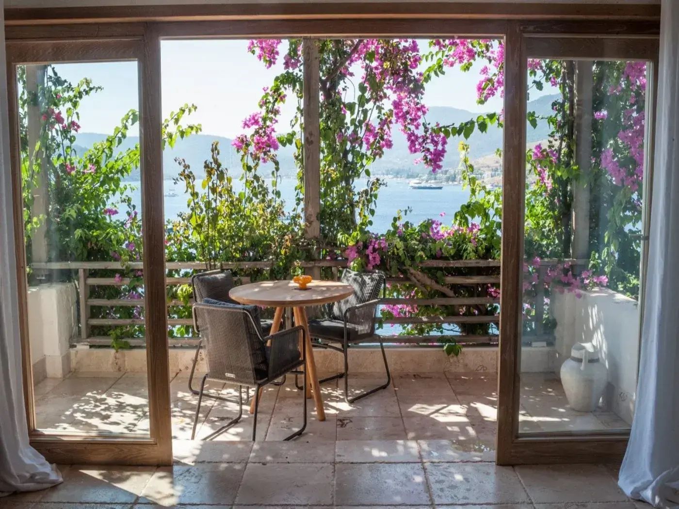 Blick von einer Terrasse mit Tisch und Stühlen über pinke Bougainvillea auf das Meer und Hügel.