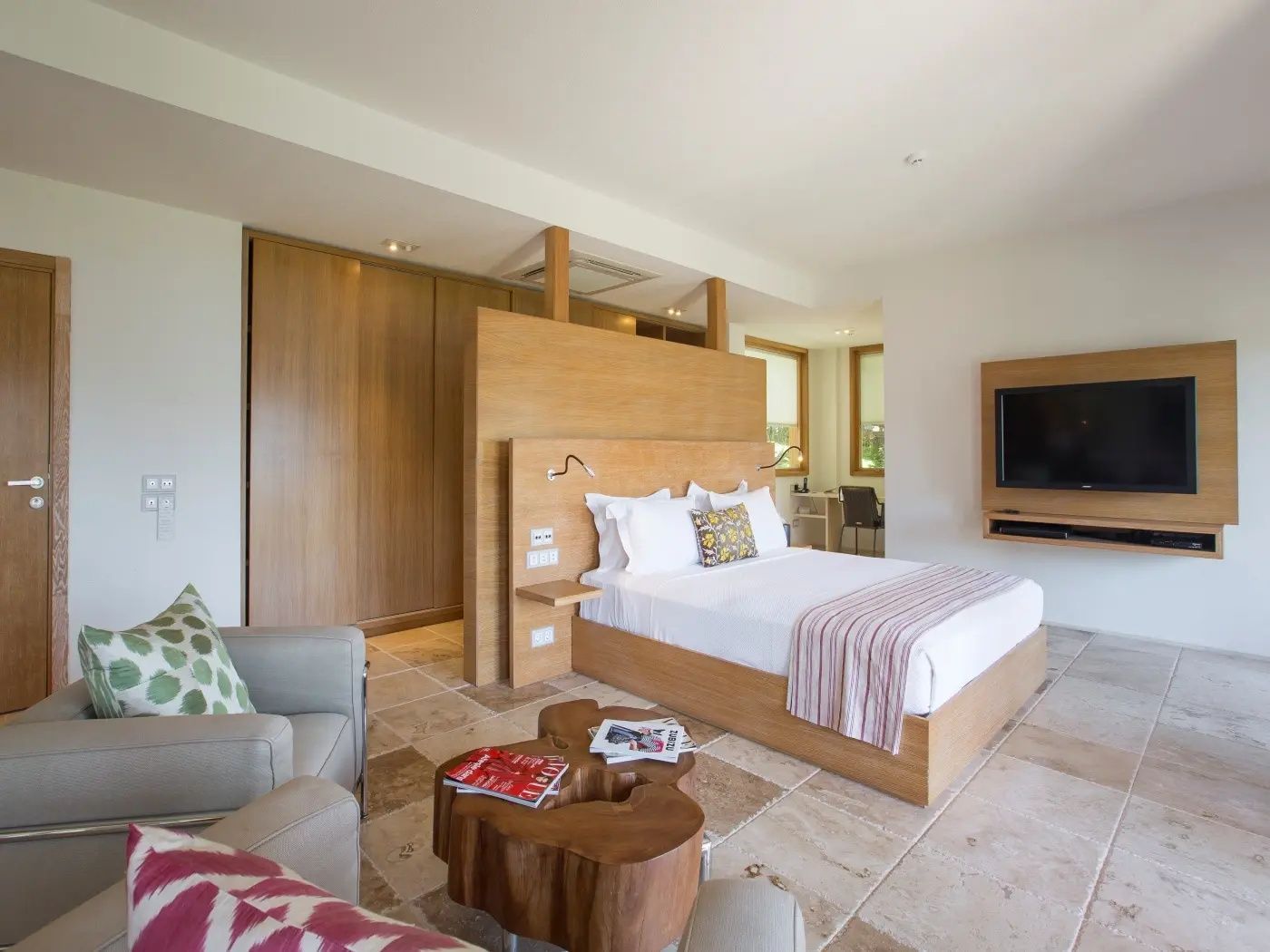 Helles Hotelzimmer mit Holzbett, Sessel, hölzernem Beistelltisch und Wandfernseher.