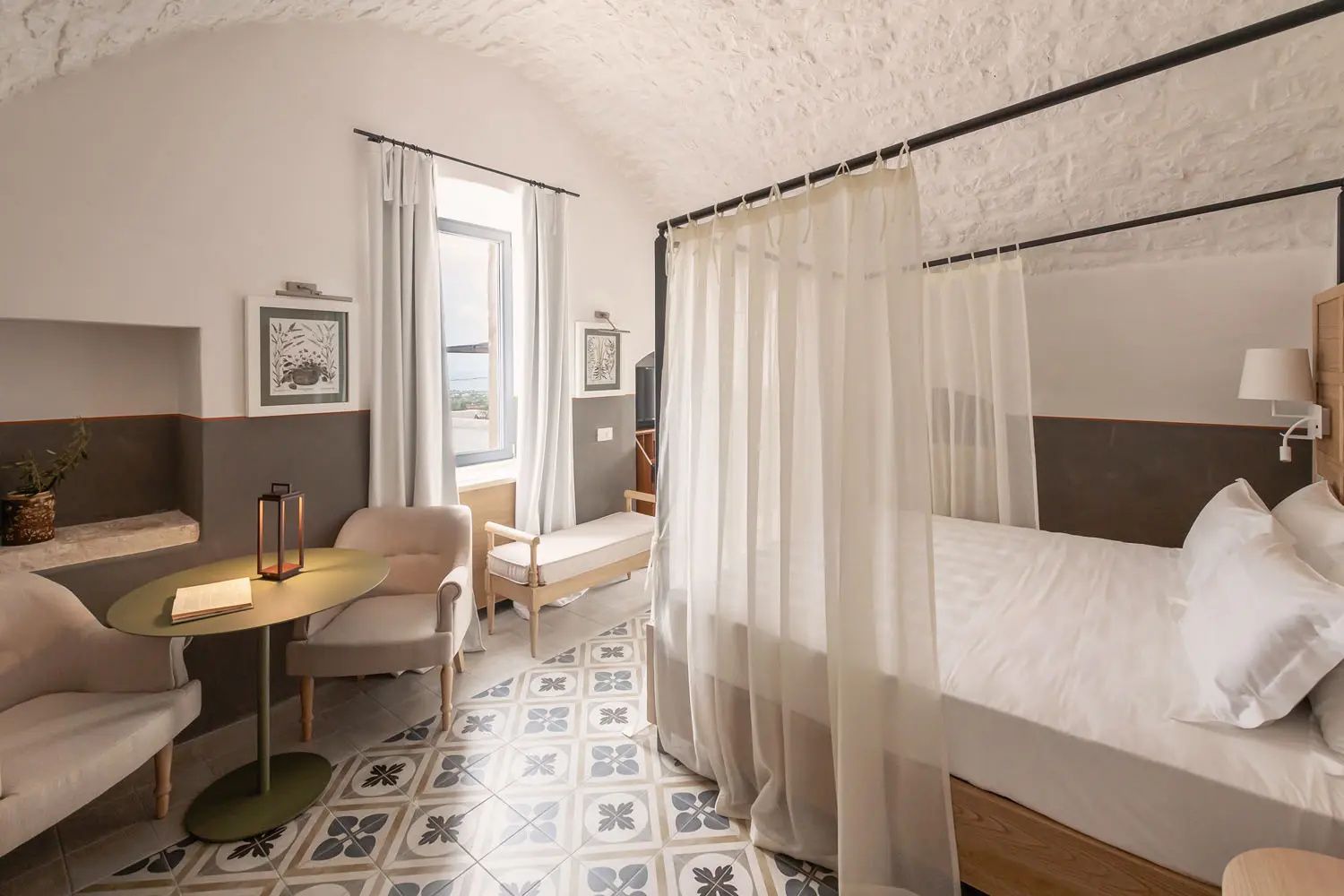 Helles Hotelzimmer mit Himmelbett, gewölbter Decke, gemustertem Fliesenboden und Sitzbereich am Fenster.