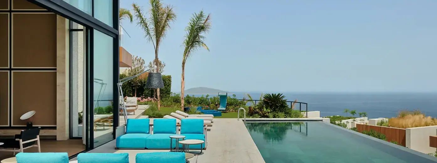 Außenpoolbereich mit blauen Sitzgelegenheiten, Liegen, Palmen und Blick auf das Meer.