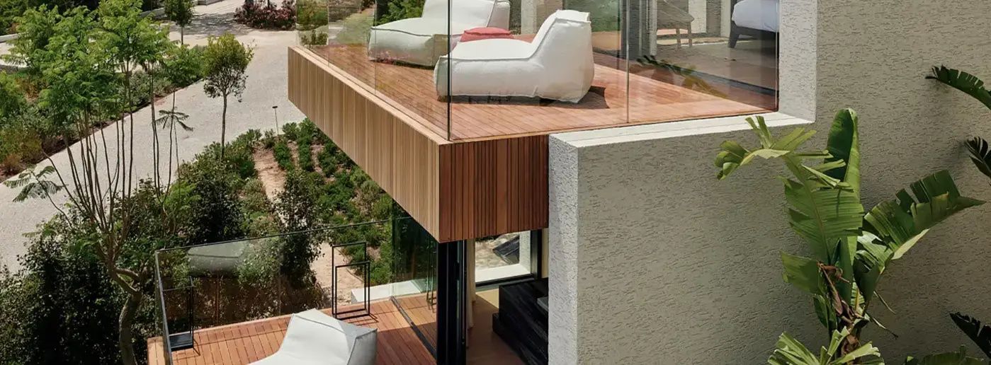 Holzterrasse mit weißen Sitzmöbeln, Glasgeländer und Blick auf die grüne Umgebung.