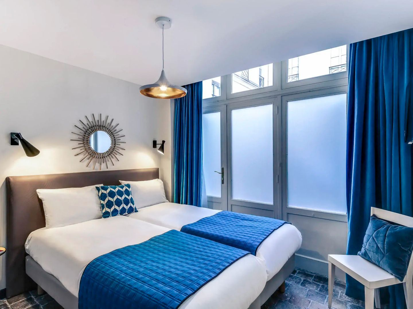 Doppelbett mit blauer Tagesdecke, blauem Kissen und Vorhängen vor einem großen Fenster.