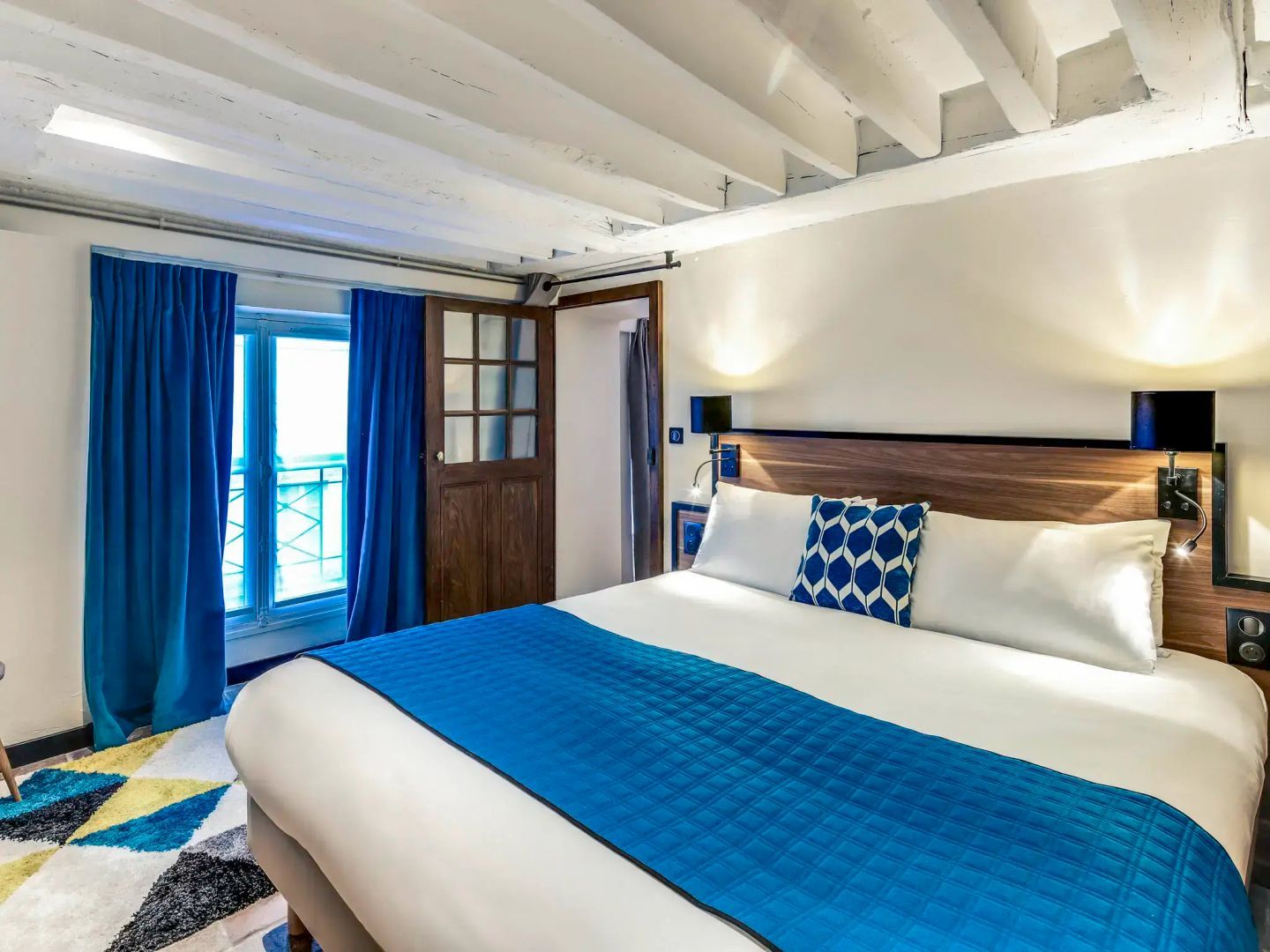 Doppelbett mit blauer Tagesdecke unter einer hellen Holzbalkendecke im Hotelzimmer.