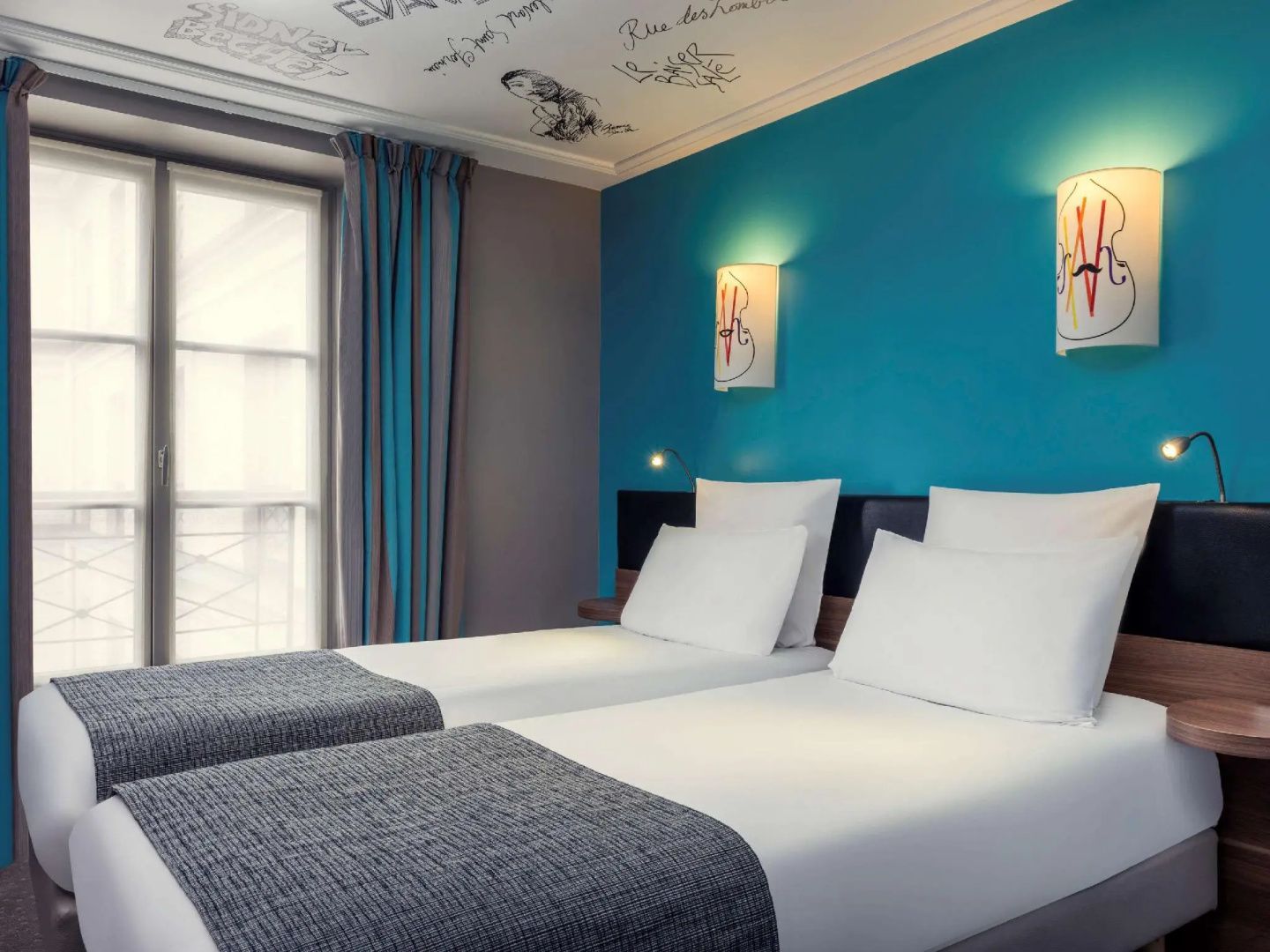 Zwei Einzelbetten vor einer blauen Wand mit Wandleuchten und verziertem Deckenmuster im Hotelzimmer.