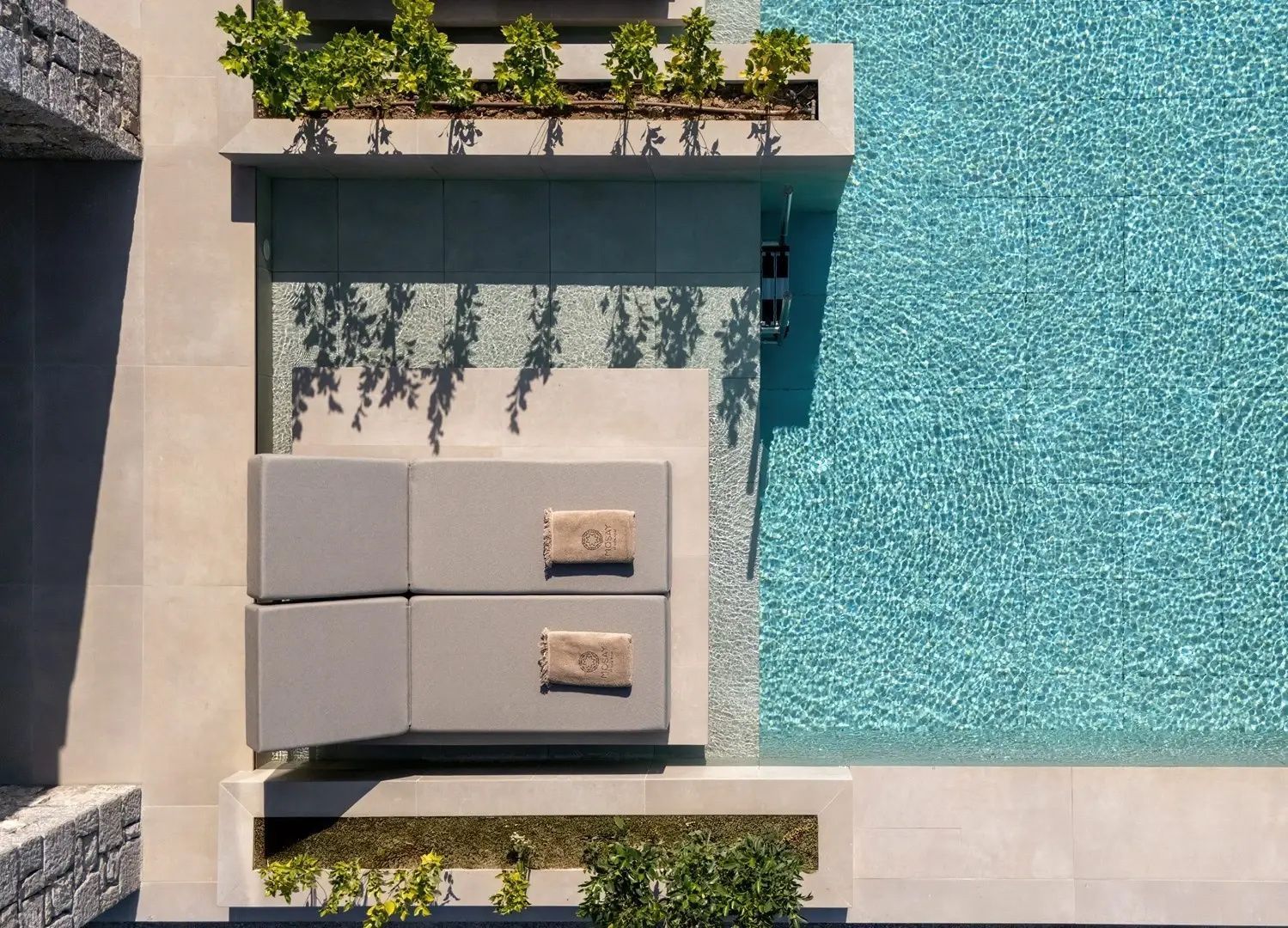 Draufsicht auf zwei Sonnenliegen direkt am Poolrand mit blauem Wasser.