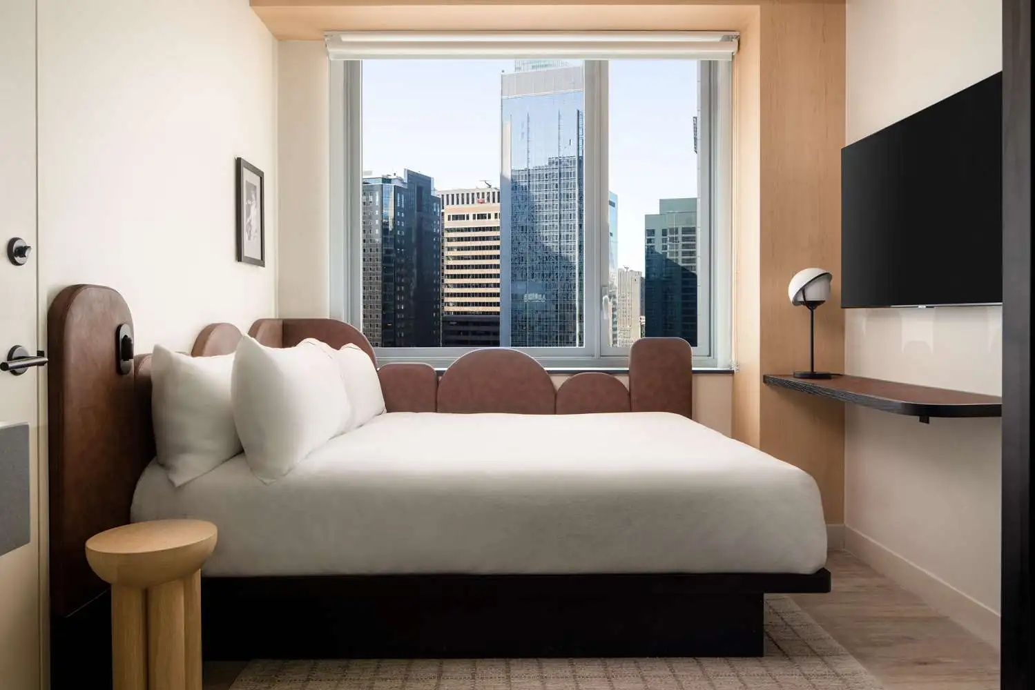 Helles Hotelzimmer mit großem Bett vor einem Fenster mit Aussicht auf die Skyline von New York.