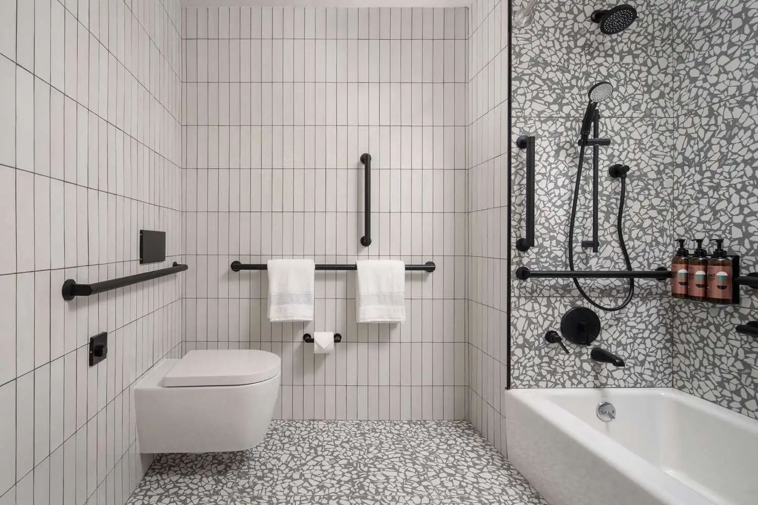 Barrierefreies Badezimmer mit Badewanne, Toilette, Haltegriffen und Mosaikboden.