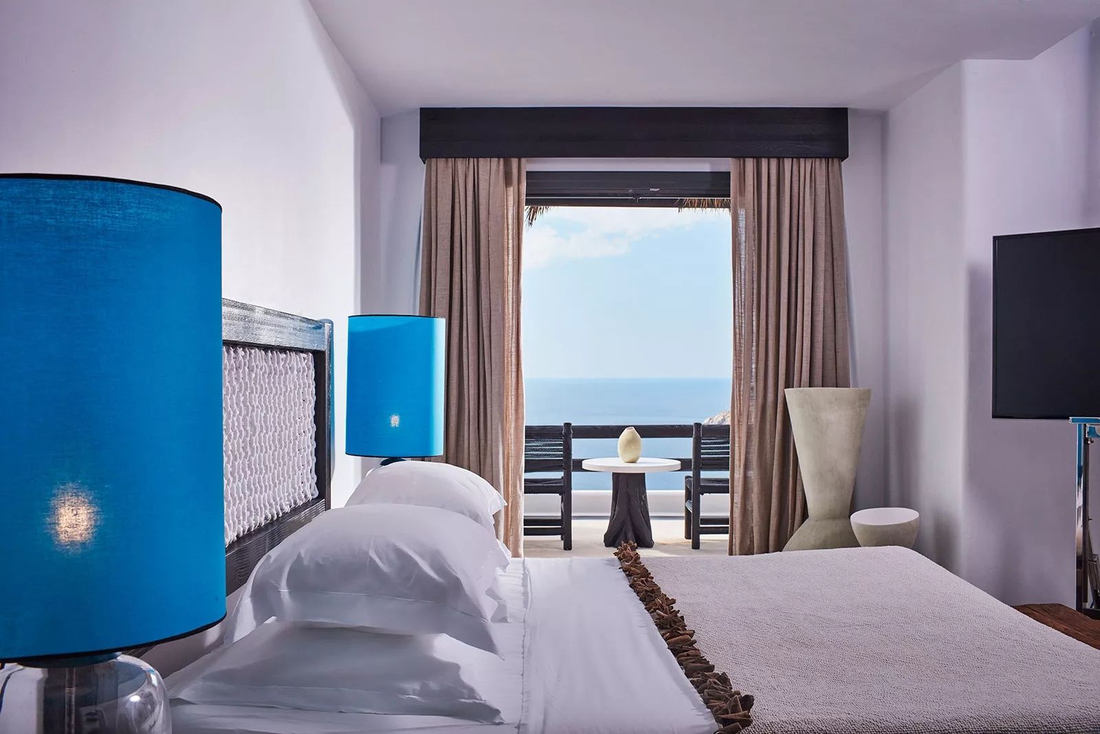 Helles Hotelzimmer mit Doppelbett, blauen Lampen und Blick auf den Balkon mit Meerblick.