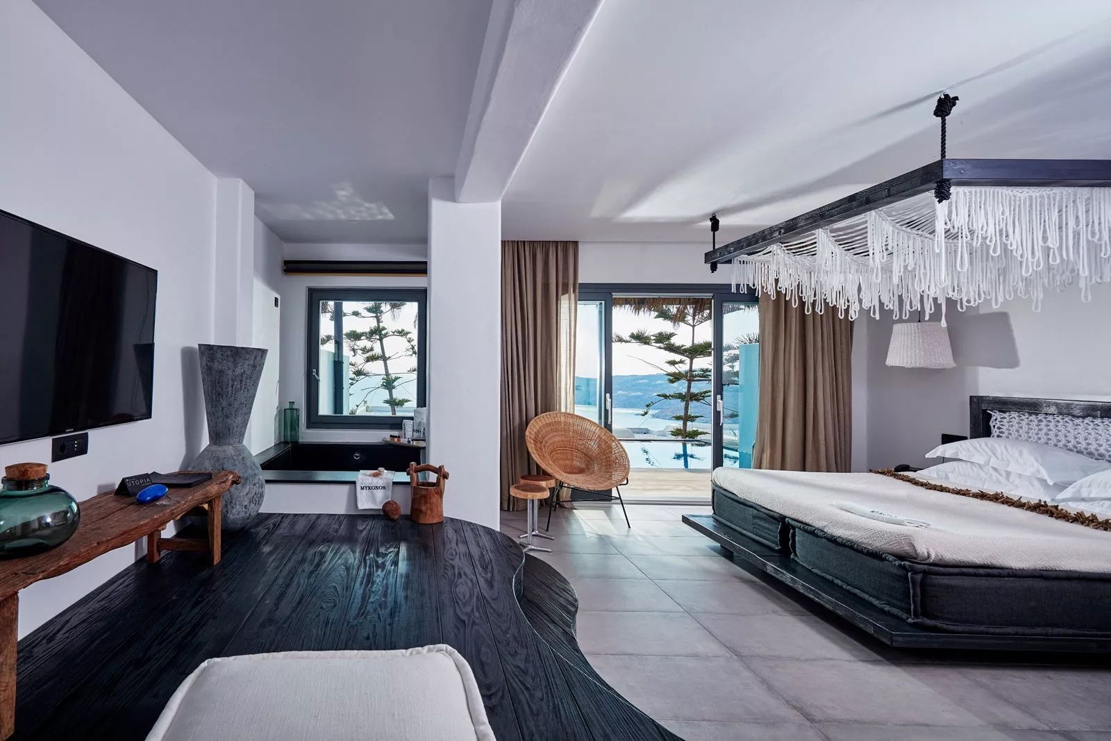 Helles Hotelzimmer mit dunklem Bett, Weidensessel und Terrasse mit Blick auf das Meer.
