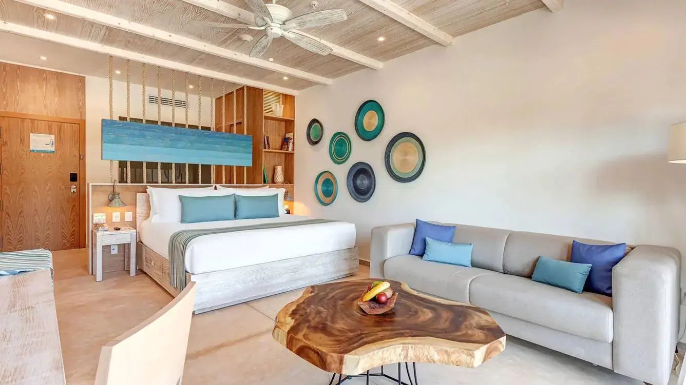 Hell gestaltetes Hotelzimmer mit grauem Sofa, Holztisch und Bett im Hintergrund.